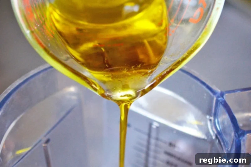 Pouring golden olive oil from a liquid measuring cup into an open blender container to begin the emulsification of Tonnato Sauce.