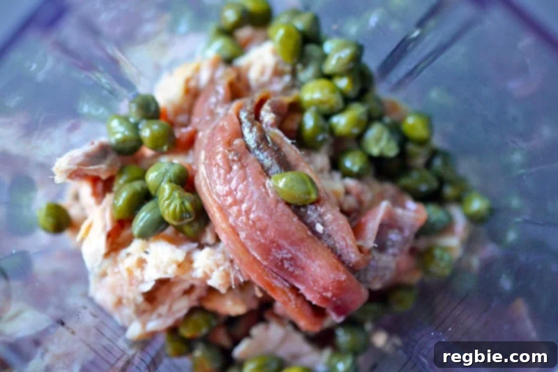 An overhead shot of the inside of a blender filled with all the foundational ingredients for Tonnato Sauce – tuna, capers, and anchovies – before blending.