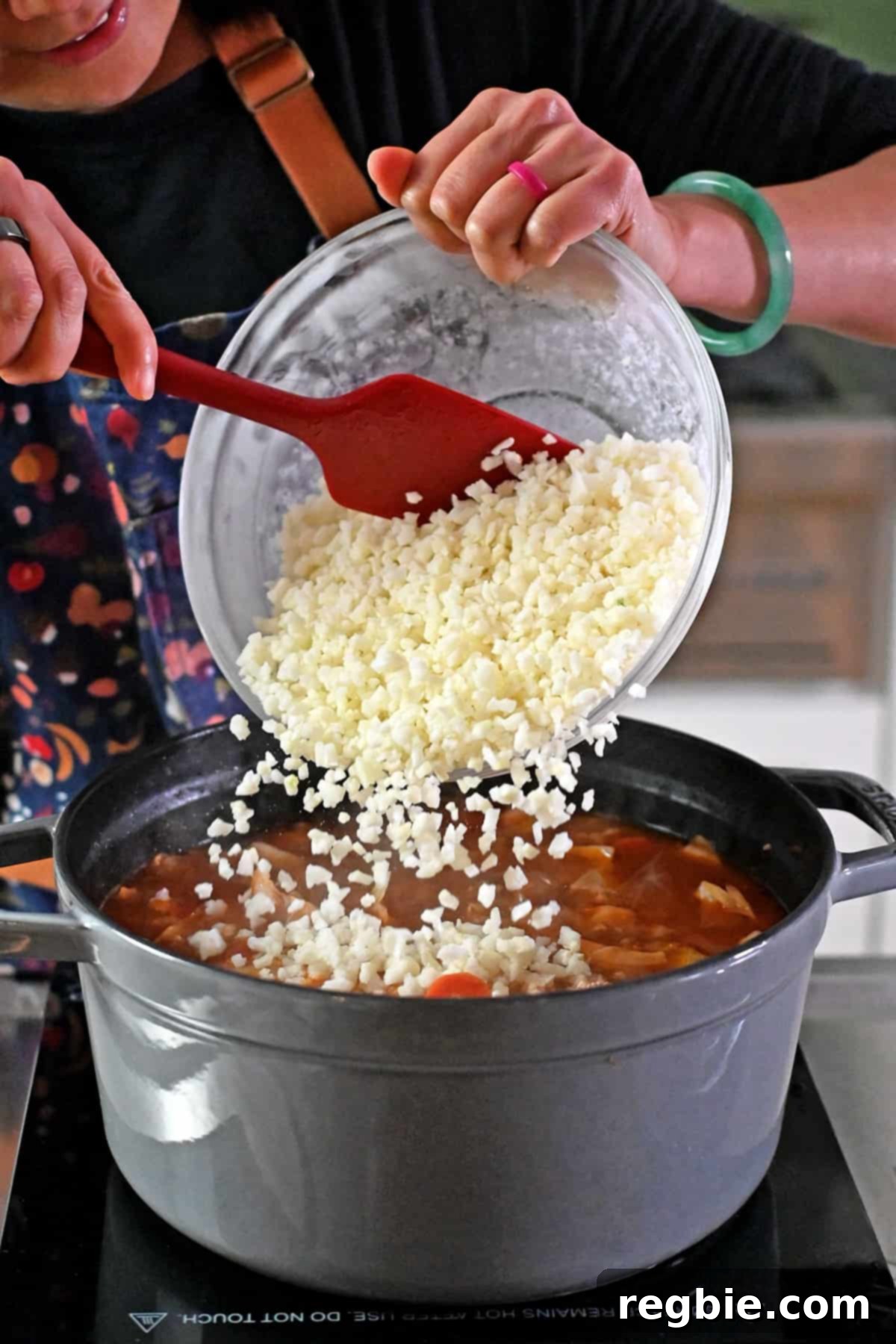 Adding a bowl of frozen riced cauliflower to a simmering pot of cabbage roll soup.