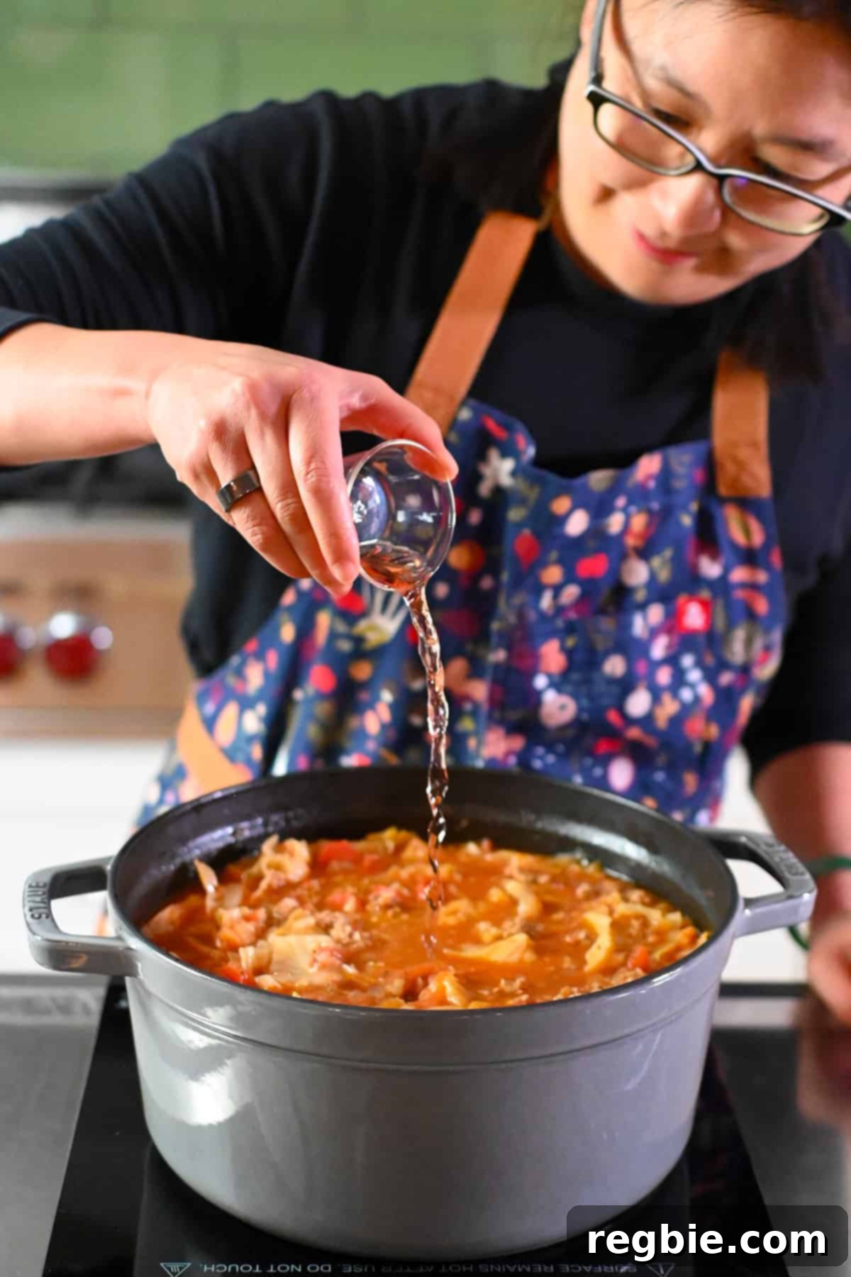 An Asian woman in glasses is adding some red wine vinegar to a gray stockpot filled with cabbage roll soup, while stirring.