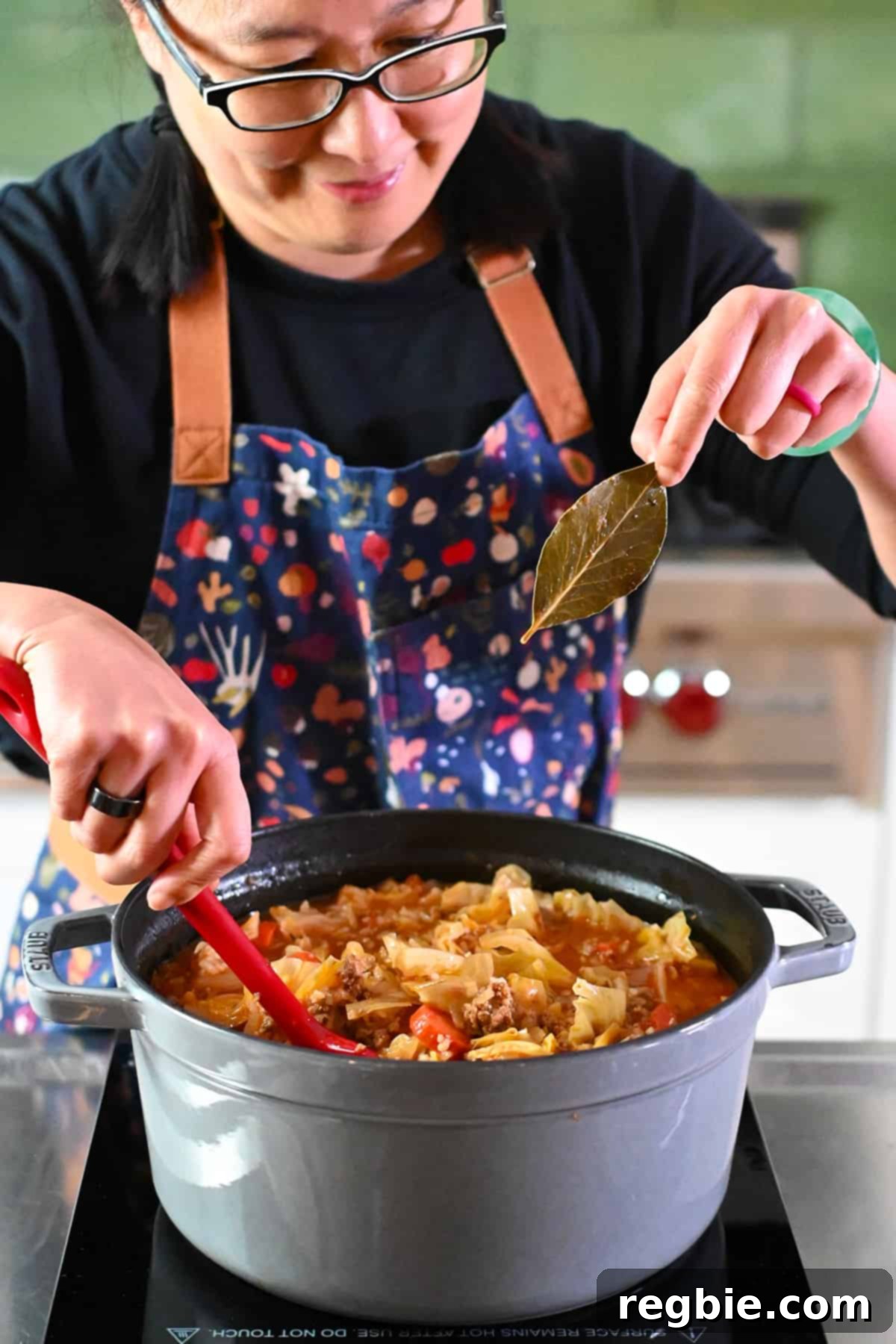 A smiling Asian woman is picking a bay leaf out of a pot of cabbage roll soup, smiling.
