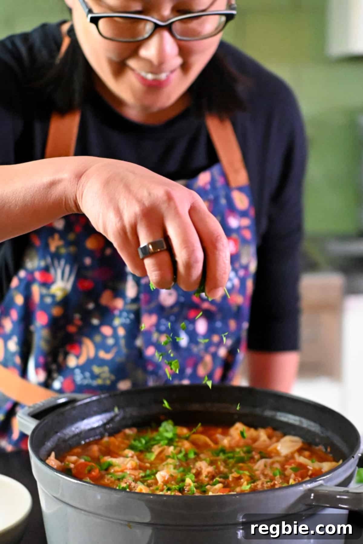 A smiling Asian woman is adding a sprinkle of minced Italian parsley to a big pot of cabbage roll soup as a garnish.
