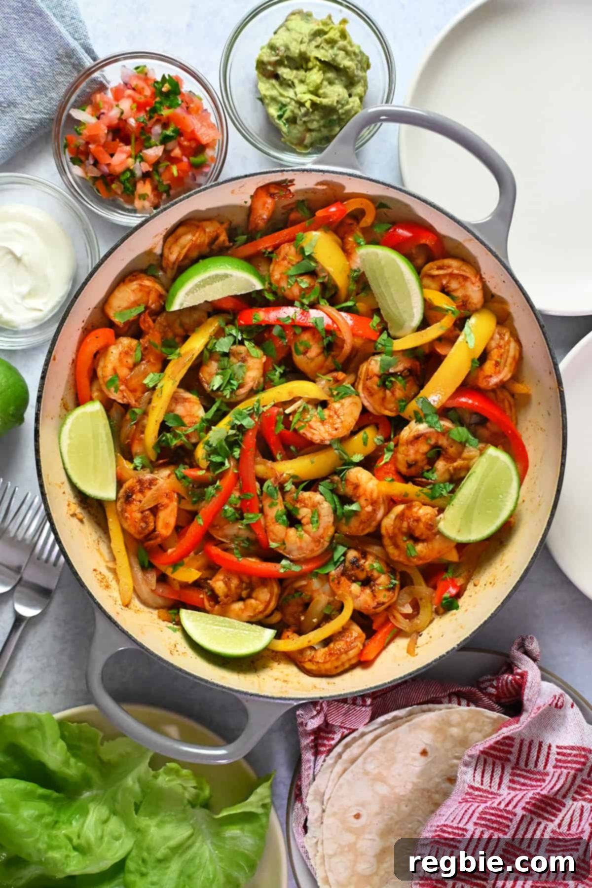 An overhead shot of a skillet filled with vibrant shrimp fajitas, artfully arranged with sides of creamy guacamole, zesty salsa, cooling sour cream, fresh lettuce leaves, and soft tortillas.