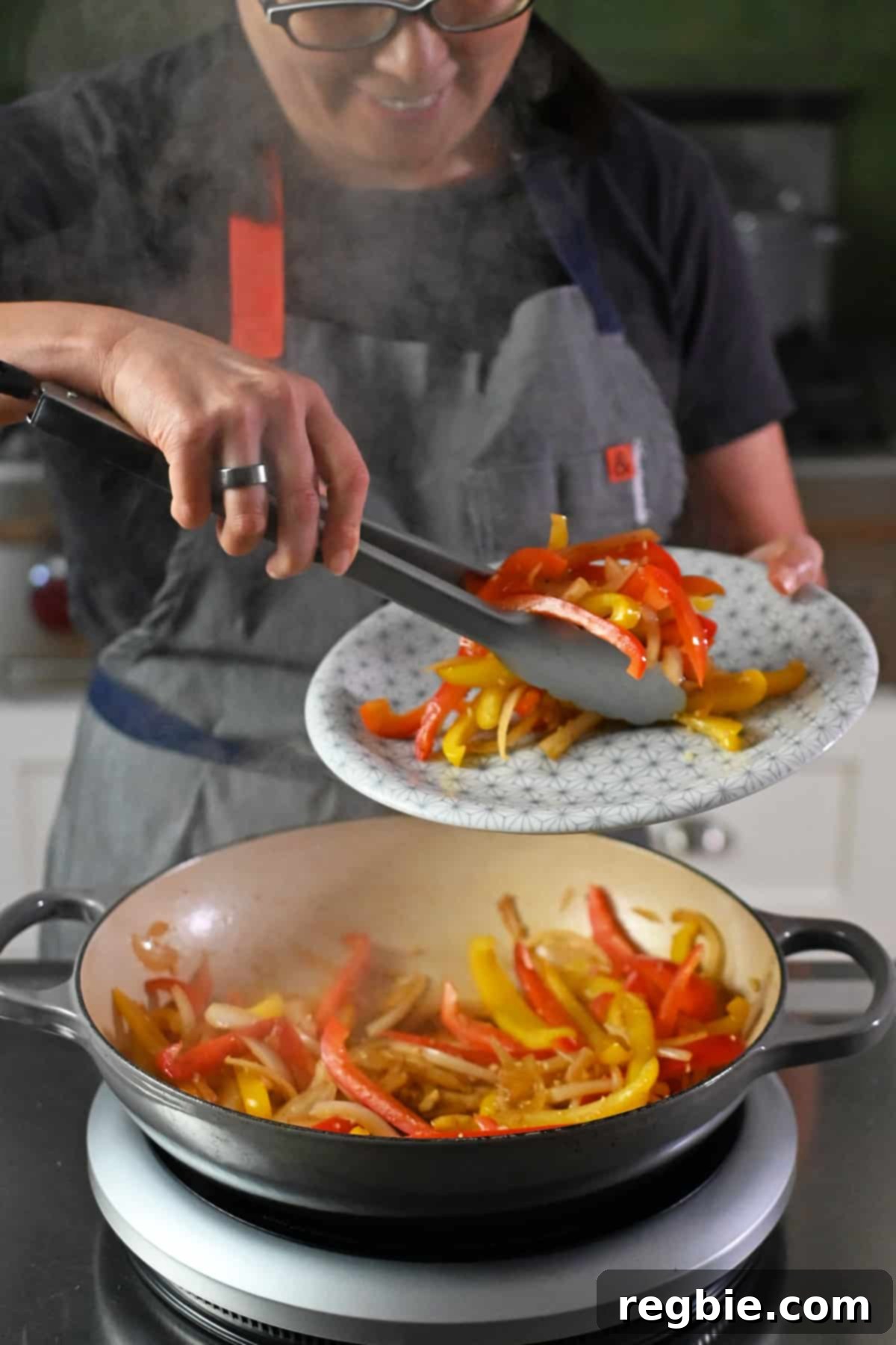 A smiling Asian woman wearing glasses, expertly transferring beautifully cooked fajita vegetables from a sizzling cast iron pan to a serving platter using a pair of tongs, ready for the next step.