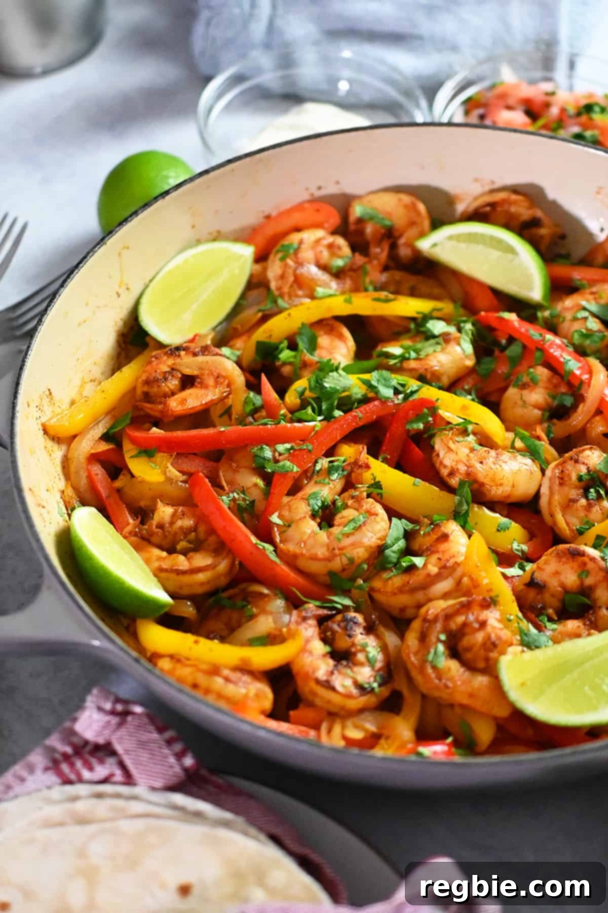 A close-up side view of a large, rustic skillet generously filled with perfectly cooked shrimp fajitas, adorned with bright lime wedges and ready to be served.