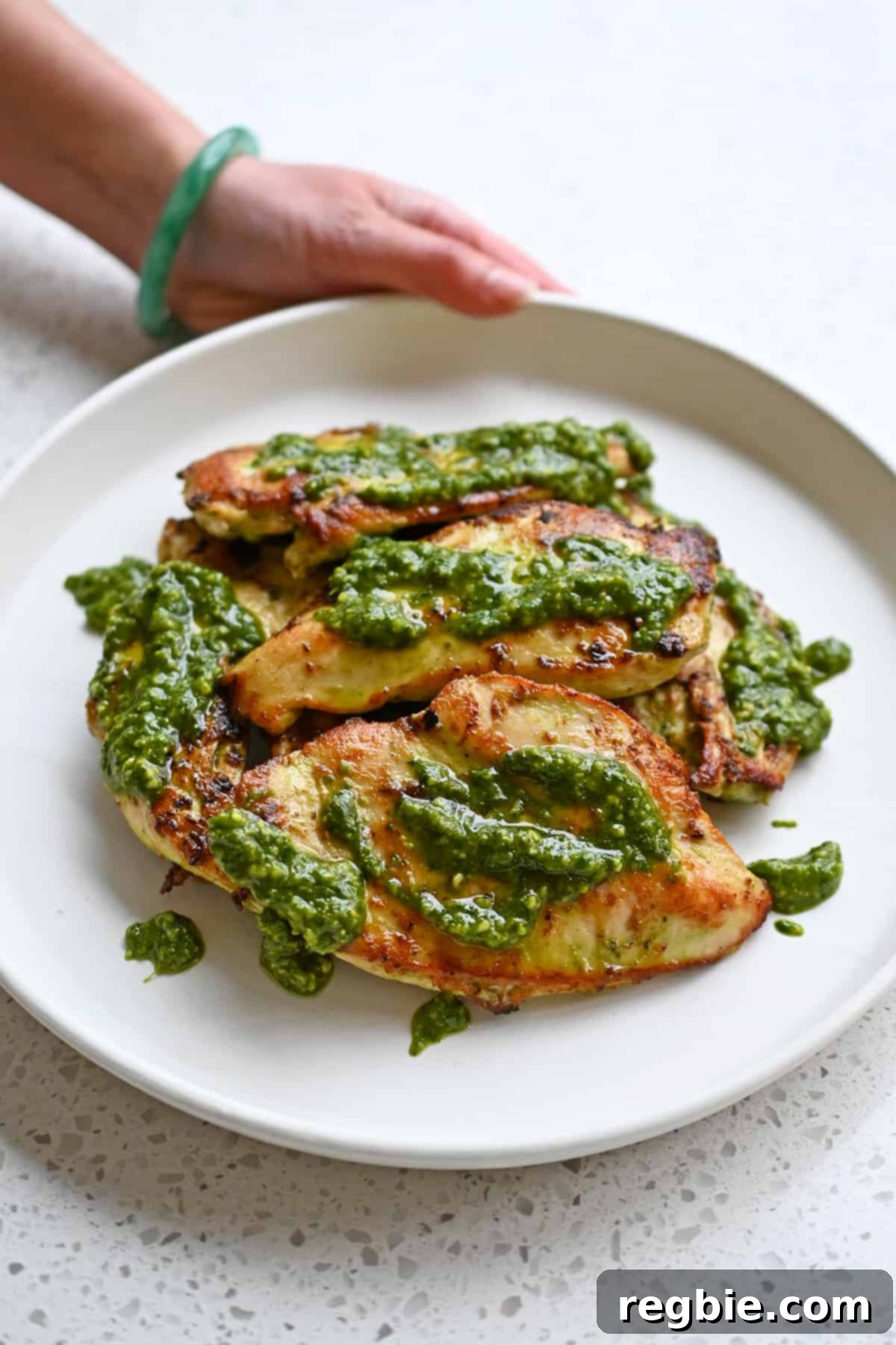 Savory Pesto Chicken 2 A hand is holding a white plate filled with pesto chicken cutlets topped with extra pesto.