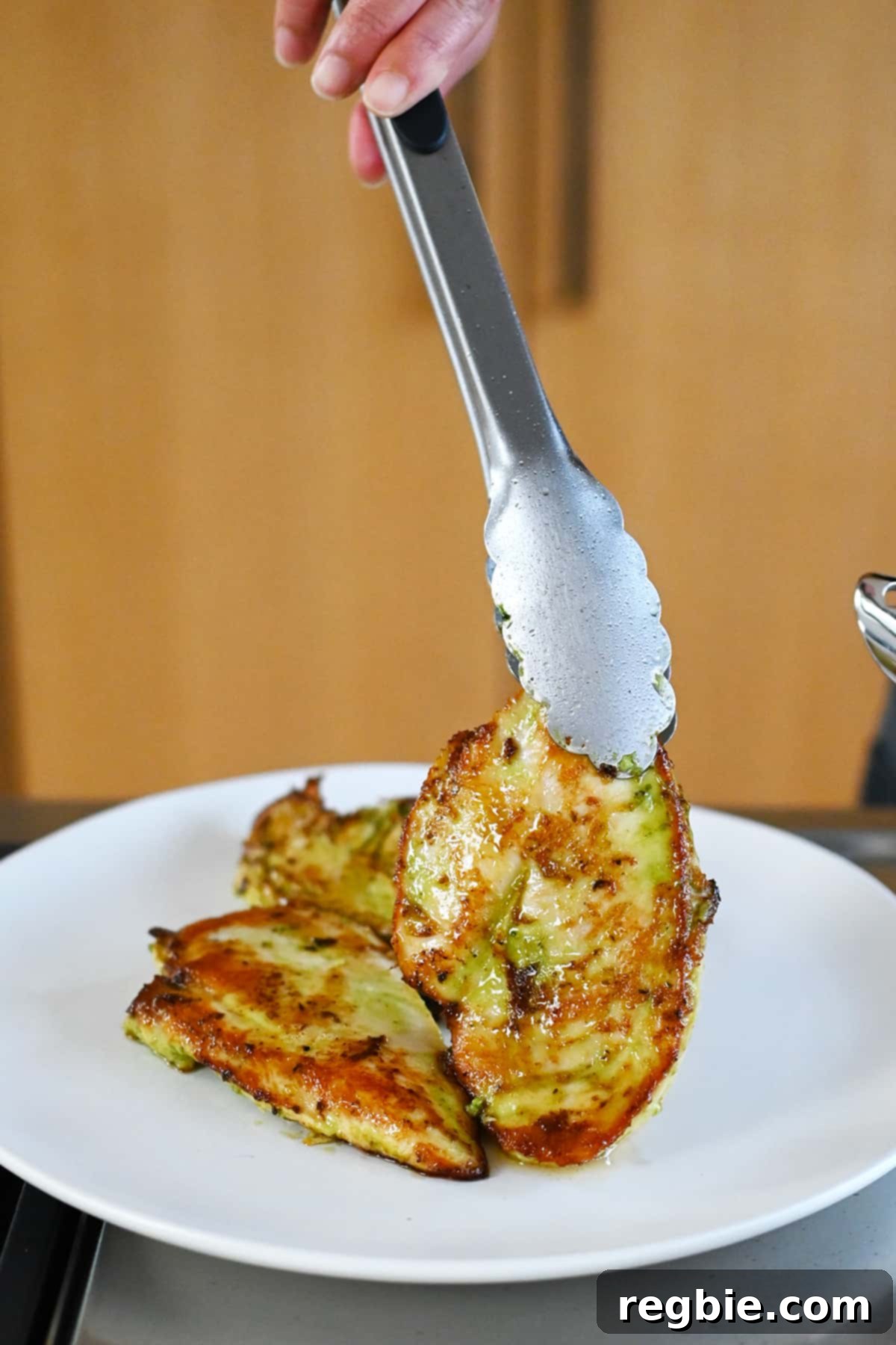Savory Pesto Chicken 12 A pair of tongs is placing a golden brown and juicy pan-seared pesto chicken cutlet onto a white plate.