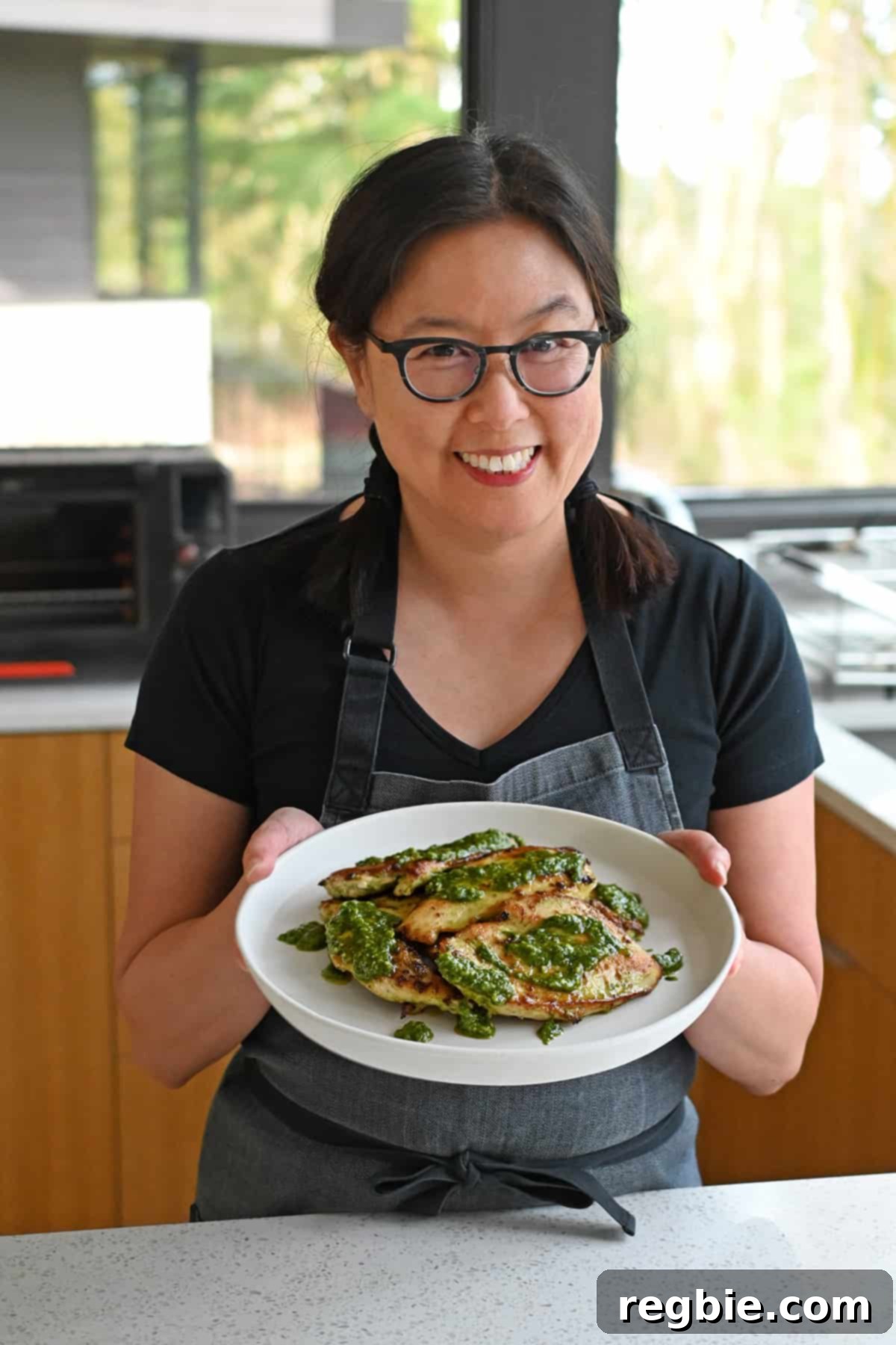 Savory Pesto Chicken 4 A smiling Asian woman wearing glasses is holding a plate filled with pesto chicken in a sunny kitchen.
