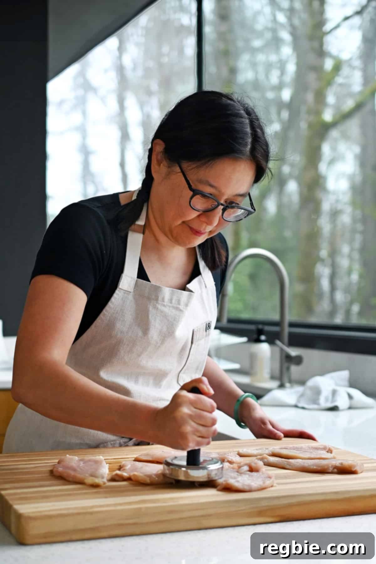 Savory Pesto Chicken 7 An Asian woman is using a meat pounder to flatten some raw chicken cutlets on a wooden cutting board before making pesto chicken.