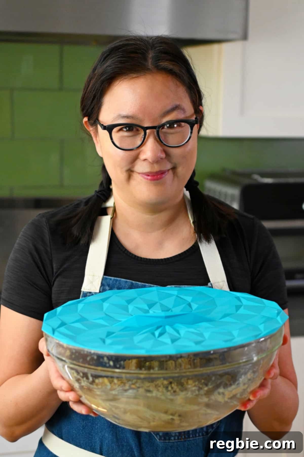 A smiling Asian woman in glasses is holding a glass mixing bowl filled with paleo banana cookie batter covered with a blue silicone lid, ready for chilling.