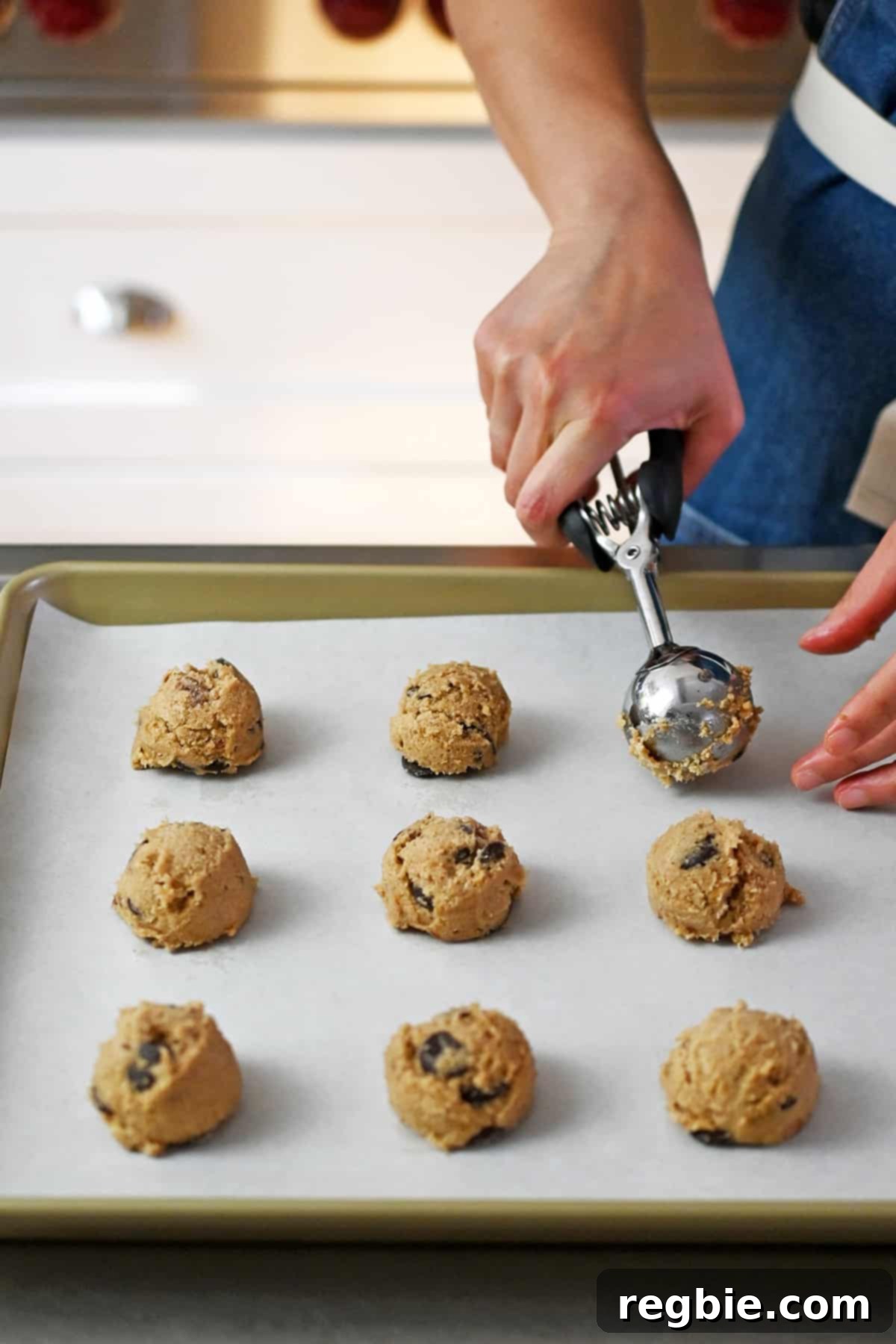 A rimmed baking sheet lined with parchment paper is covered with raw banana cookie dough balls being portioned out with a medium ice cream scooper, ready for the oven.