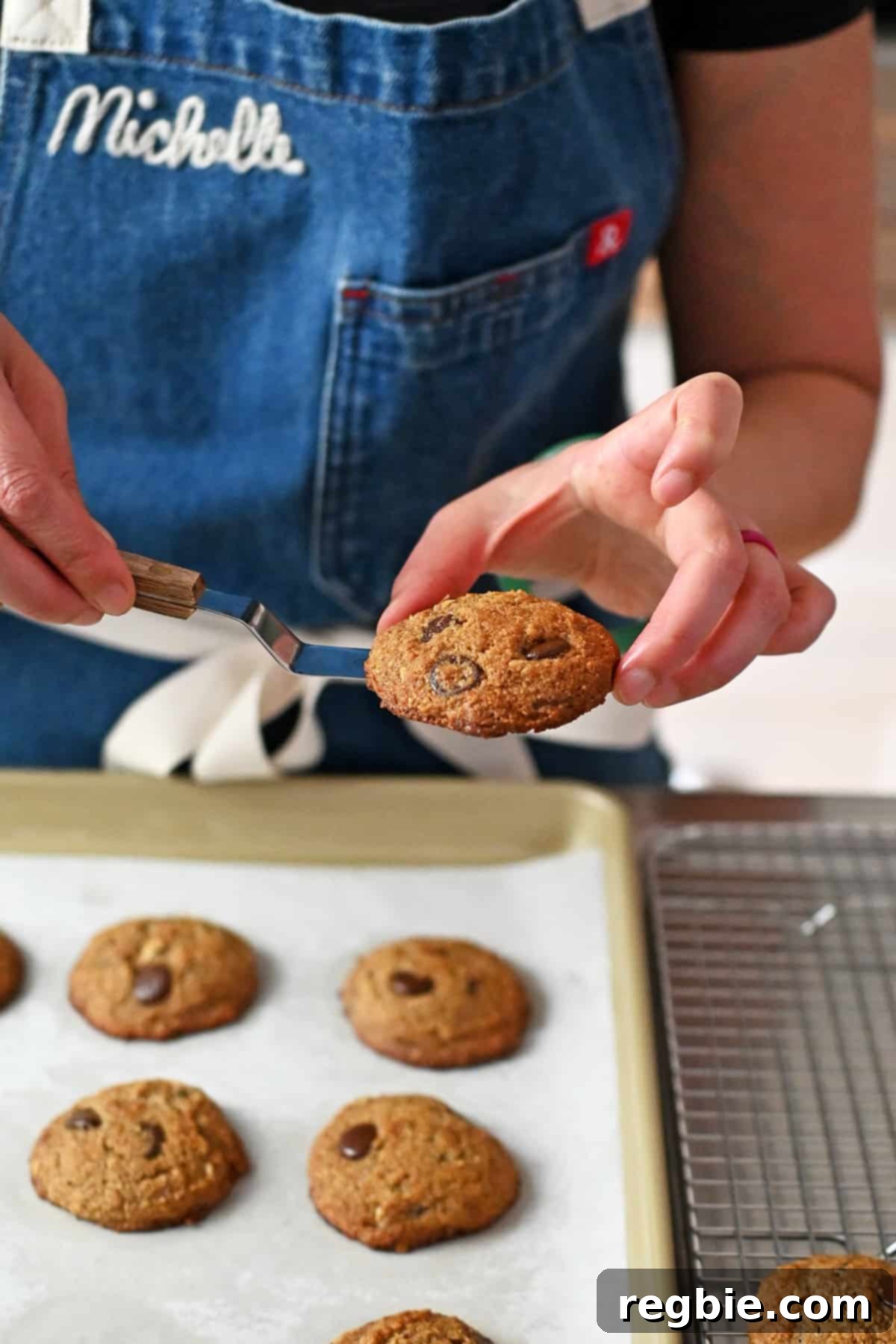 A person in a blue apron is using a small offset spatula to transfer freshly baked paleo banana cookies from a baking pan to a wire cooling rack, demonstrating the careful handling of warm cookies.