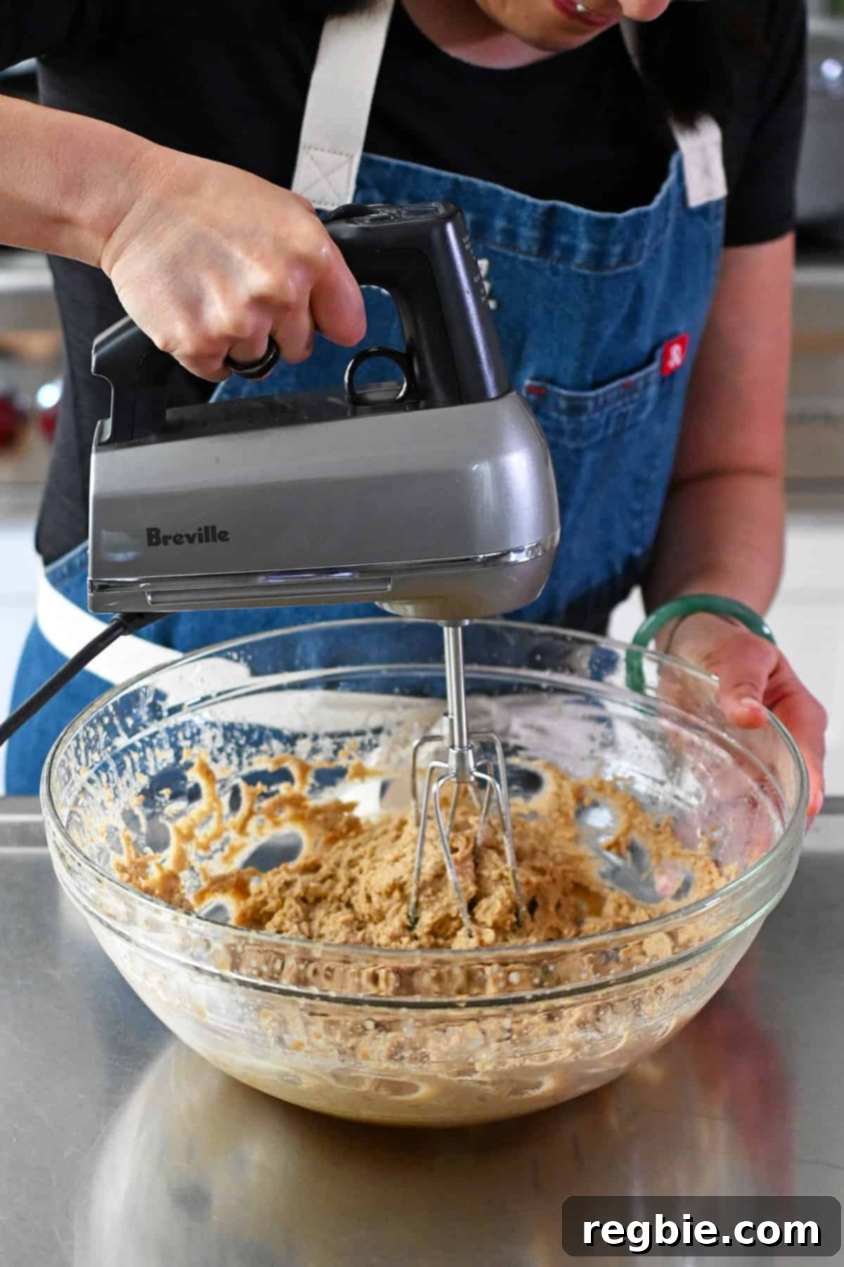 A person in a blue apron is using a hand mixer to combine the batter for paleo banana cookies in a glass mixing bowl, ensuring all ingredients are well integrated.