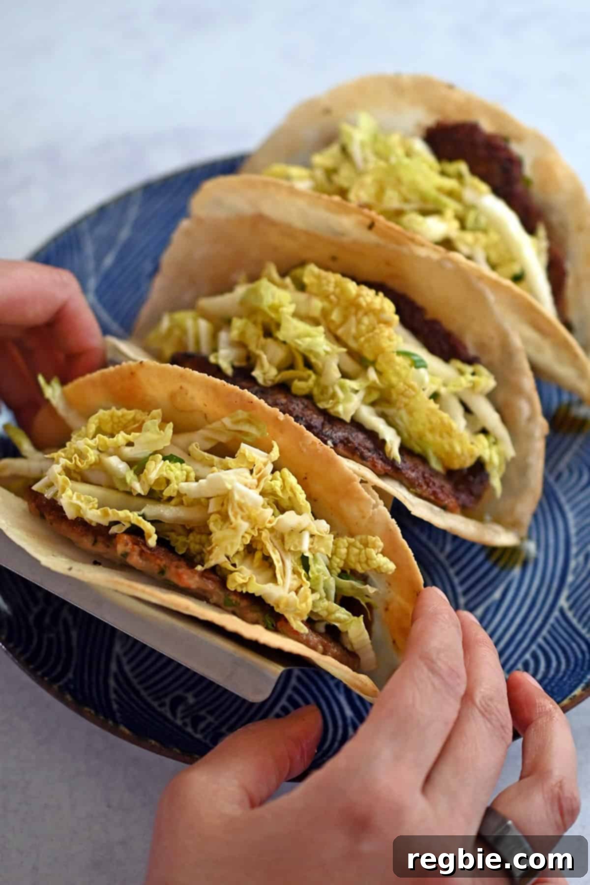 Two hands are carefully lifting a golden-brown smash dumpling taco from a vibrant blue plate, which holds two other equally tempting tacos, ready to be devoured.