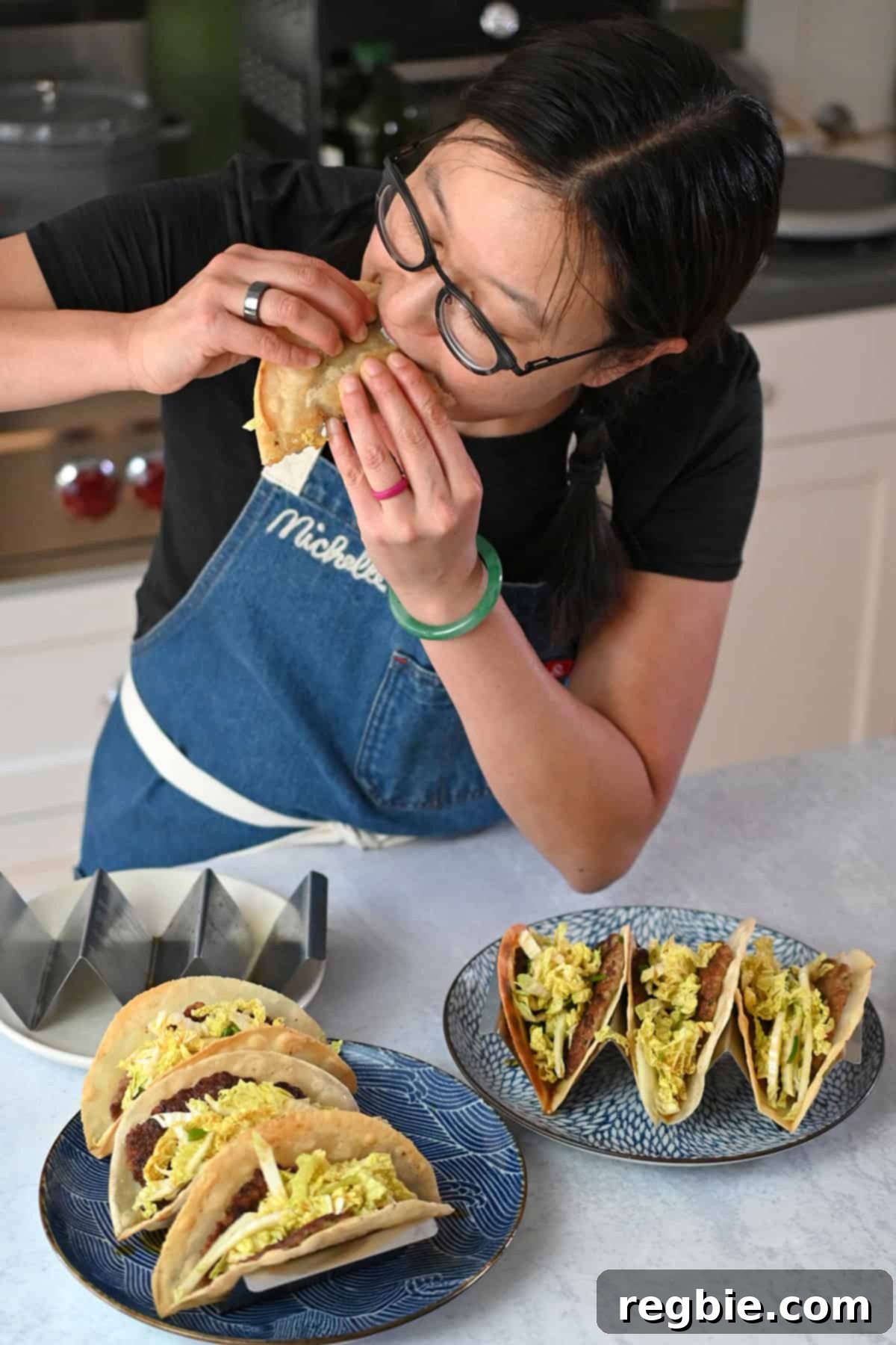 An Asian woman enthusiastically takes a bite out of a delicious smash dumpling taco, with two plates full of tacos visible in front of her.