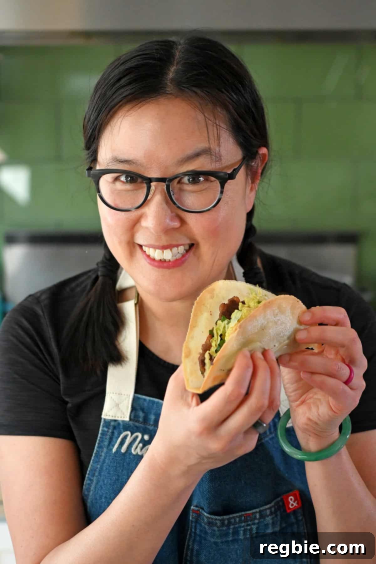 A radiant Asian woman, smiling brightly, holds up a freshly made smash dumpling taco, showcasing its enticing filling and perfectly cooked tortilla.