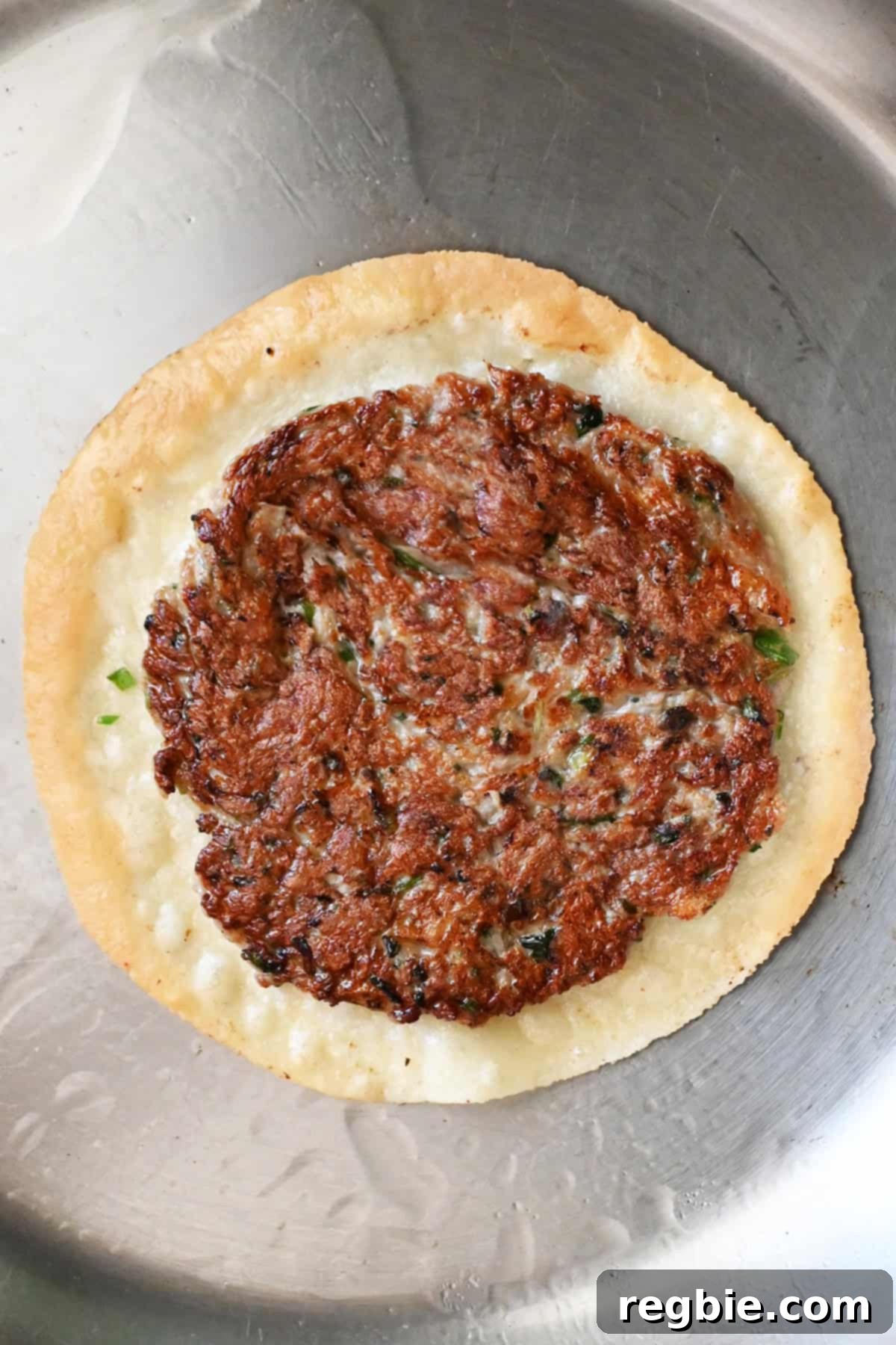 A close-up, overhead shot captures a smash dumpling taco in the pan. The meat filling is perfectly golden brown, and the tortilla beneath it is just beginning to cook to a delicate crisp.