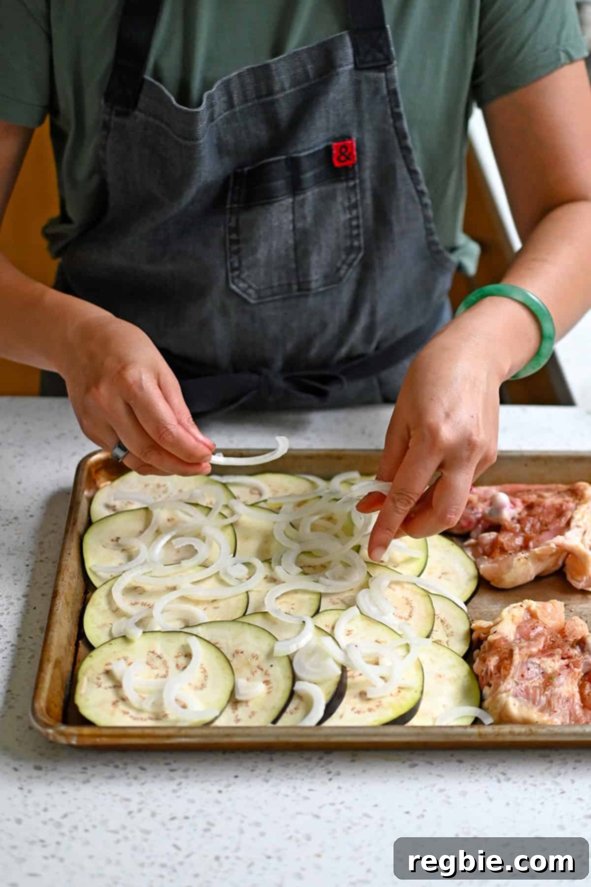 Adding thinly sliced raw onion on top of thinly sliced eggplant on a rimmed baking sheet with seasoned raw chicken thighs.