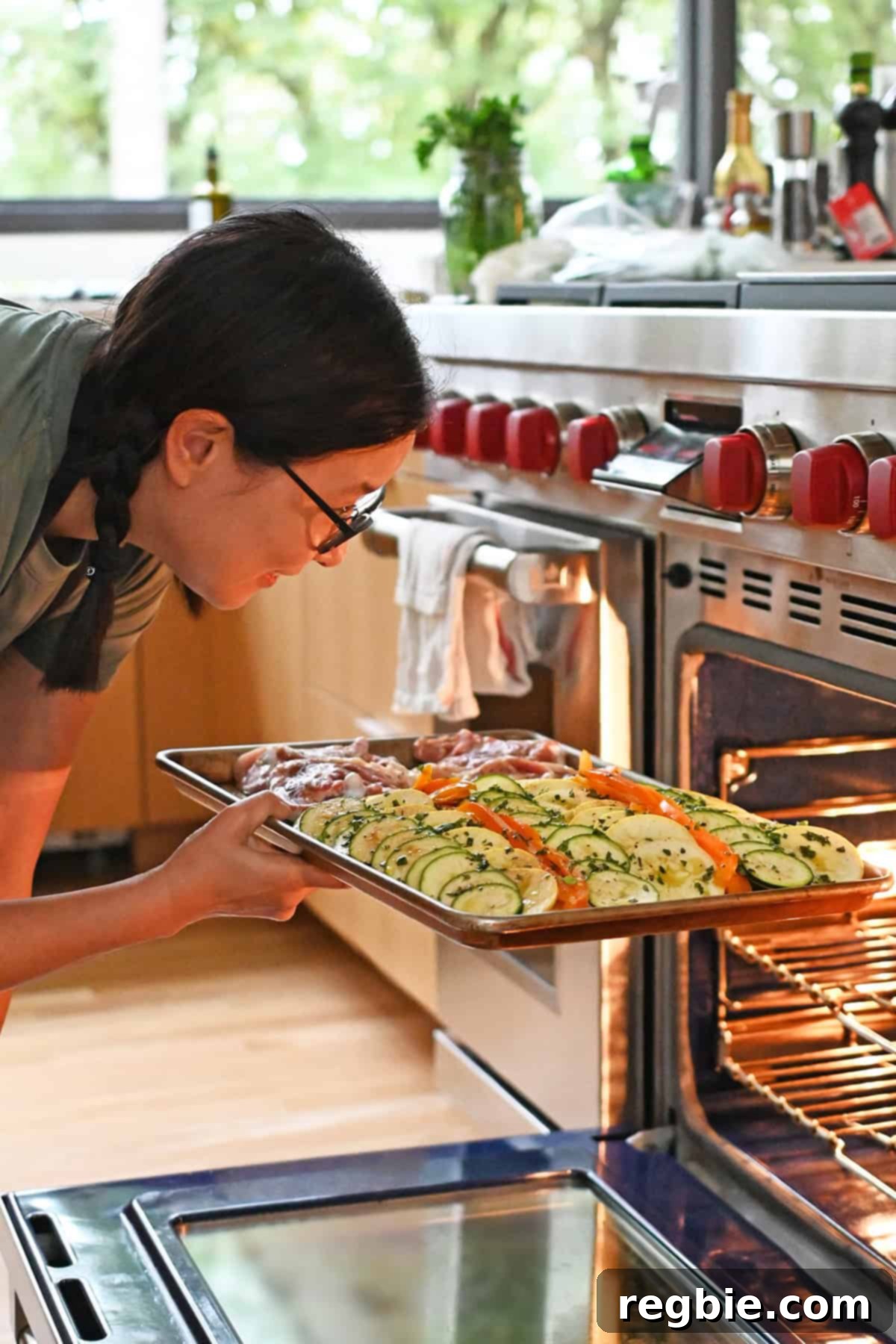 An Asian woman in glasses is putting a sheet pan filled with ratatouille vegetables and chicken thighs into the oven.