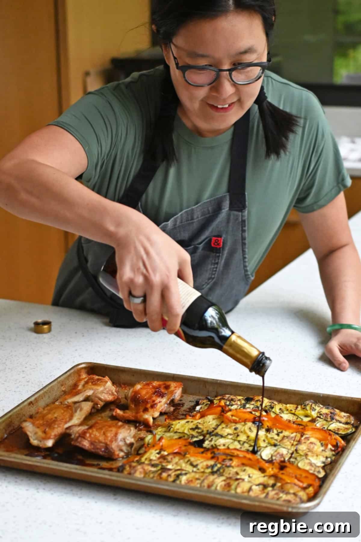 An Asian woman is drizzling aged balsamic vinegar on a sheet pan with ratatouille vegetables and golden brown chicken thighs.