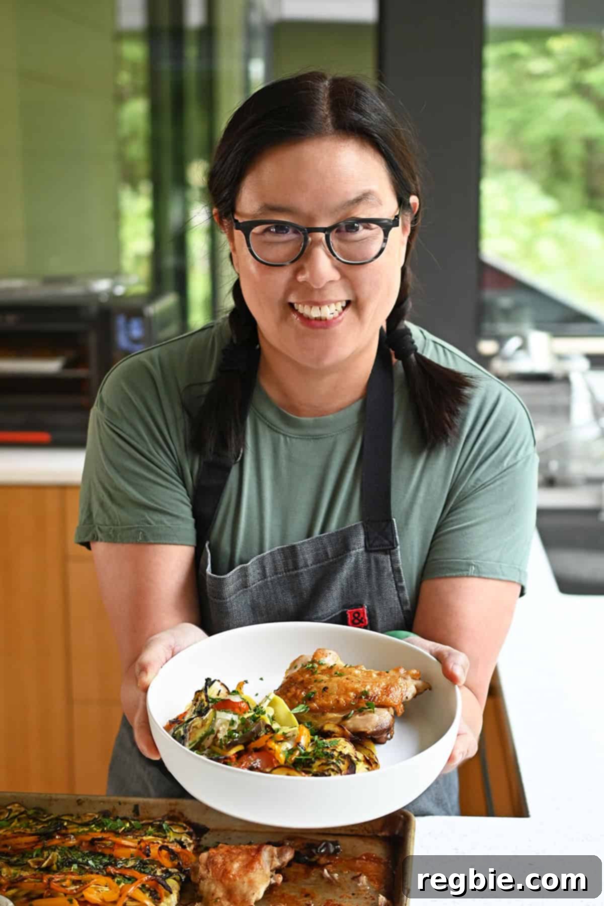A smiling Asian woman is holding a plate filled with roasted chicken thighs and ratatouille over a sheet pan filled with chicken and veggies.