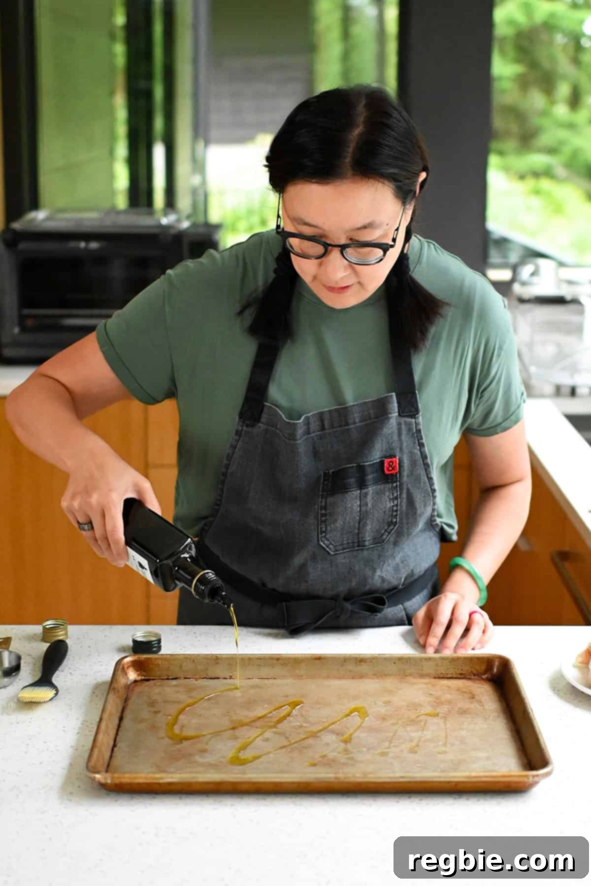 An Asian woman in glasses is pouring some extra virgin olive oil on a rimmed baking sheet.
