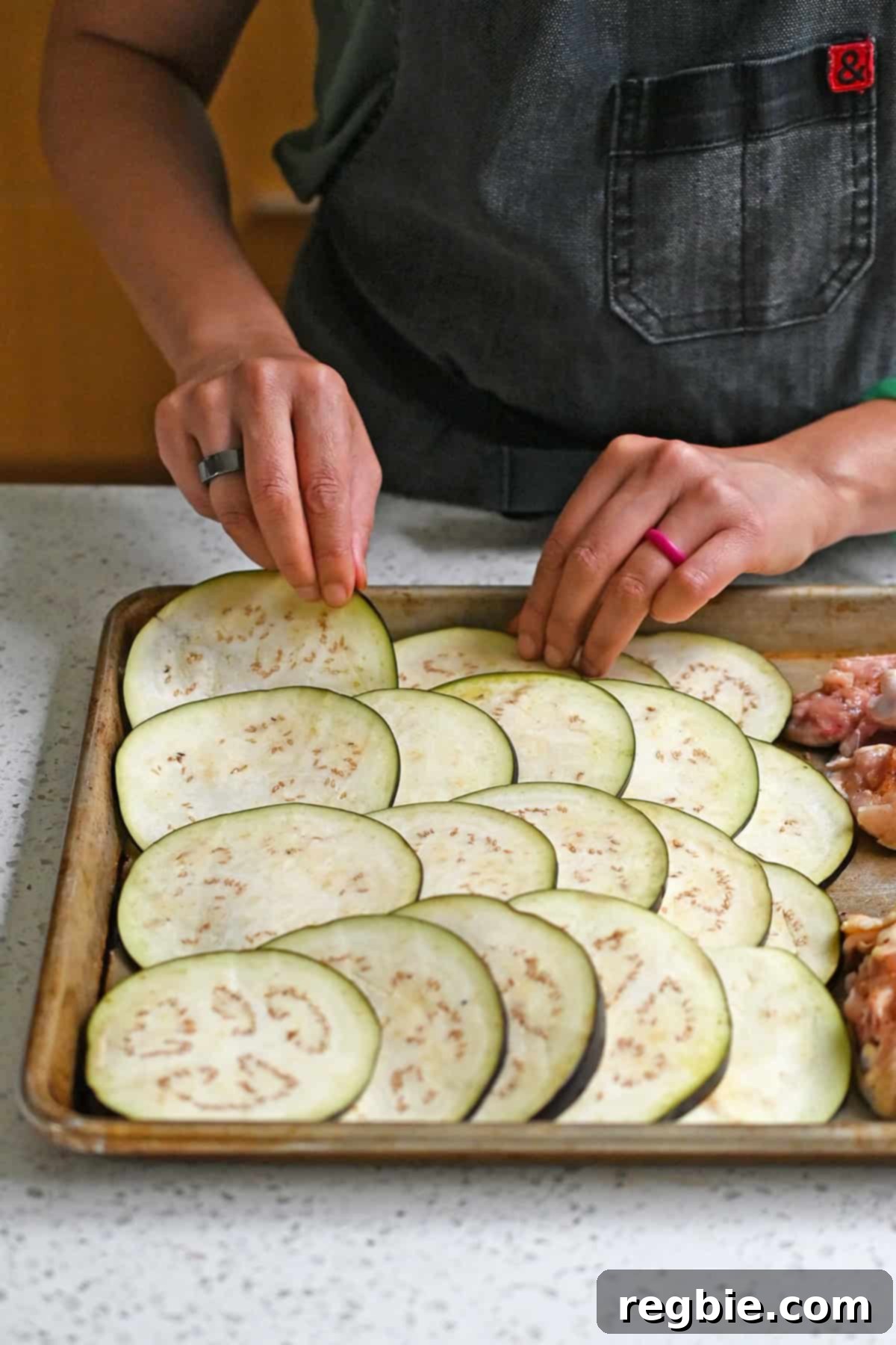 Adding a layer of thinly sliced eggplant on half a rimmed baking sheet that already has seasoned chicken thighs on it.