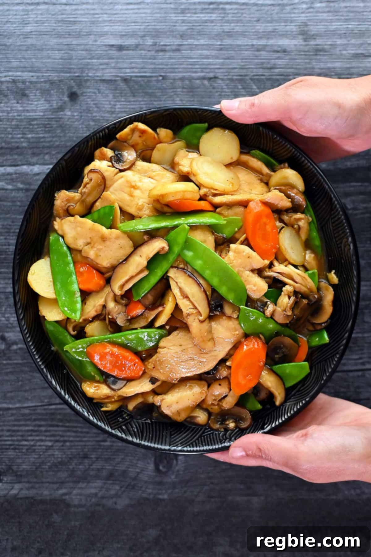 Cantonese Chicken and Mushroom Medley 2 An overhead shot of two hands holding a black porcelain bowl filled with homemade stir fried chicken and mushrooms, moo goo gai pan.