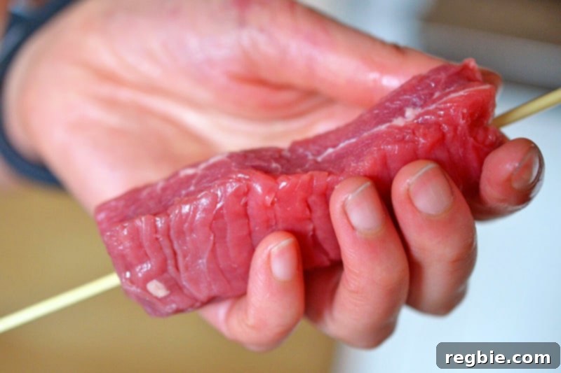 A hand is holding a piece of flank steak that has been skewered by a soaked bamboo skewer.