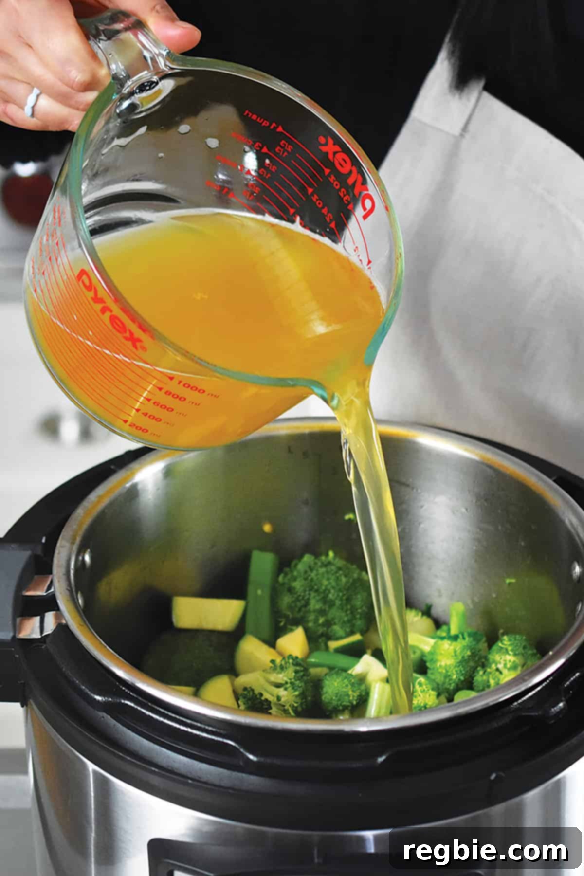 Chicken broth being poured into the Instant Pot over a colorful mix of broccoli, green onions, zucchini, and apple, ready for pressure cooking.