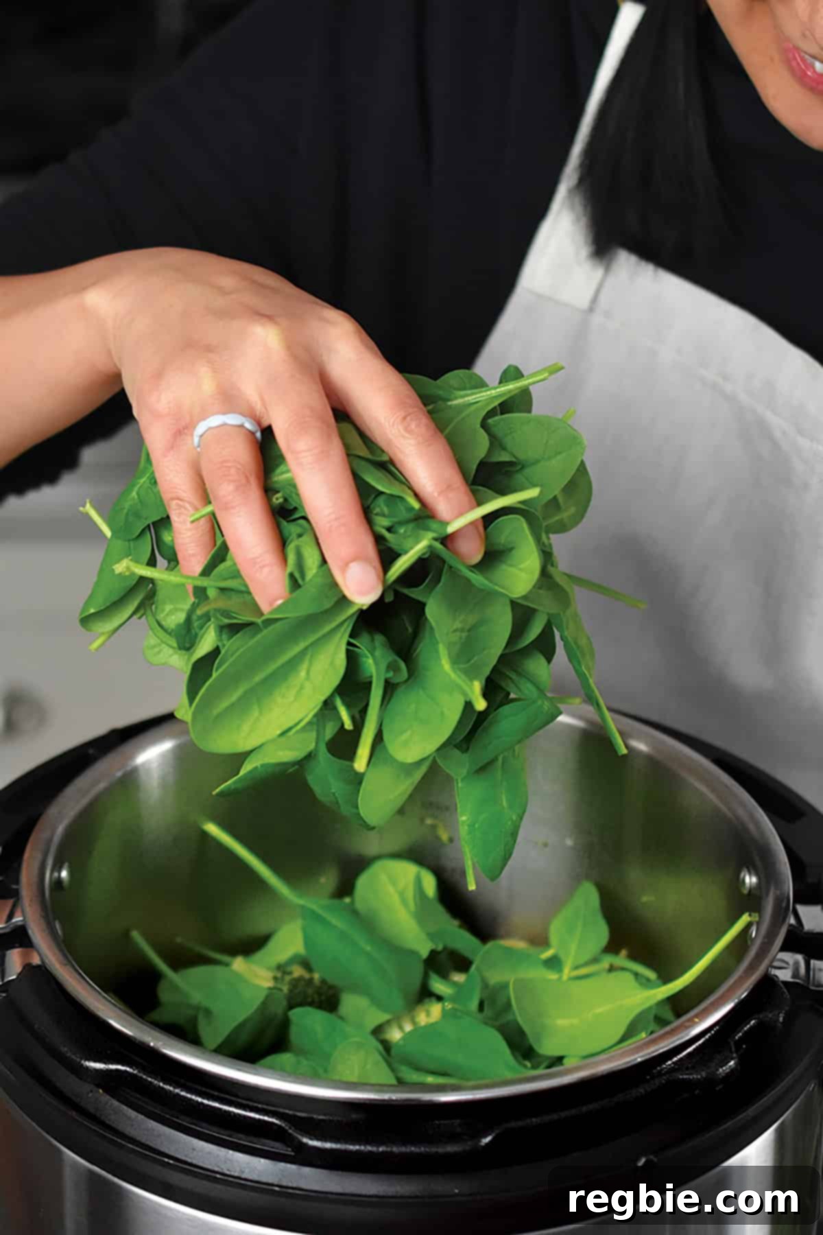 A hand adding fresh baby spinach leaves to the open Instant Pot, where the cooked green soup ingredients are still hot, ready for a quick wilt.