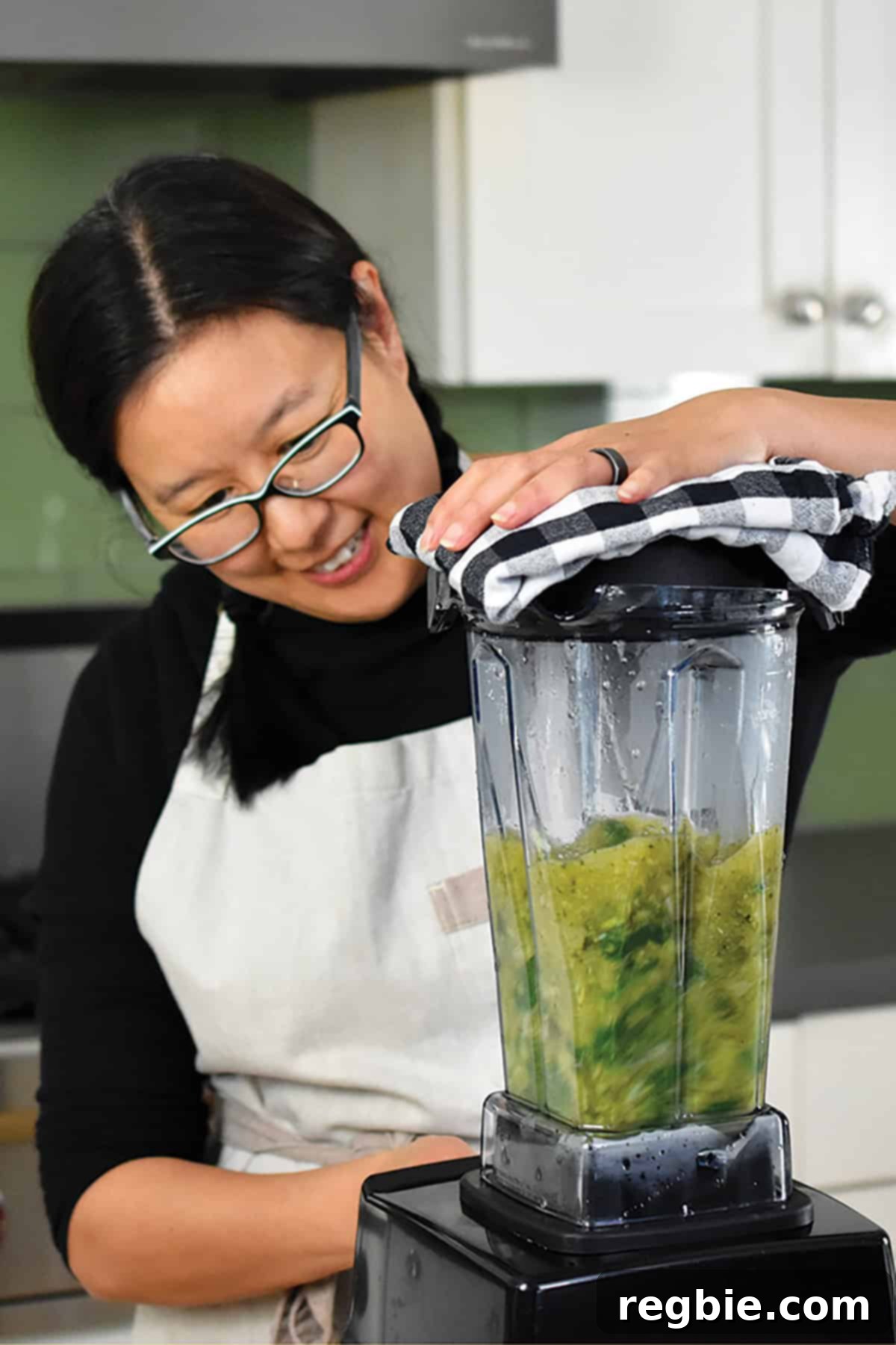 A smiling woman is blending the hot green soup in a Vitamix, demonstrating safe blending practices for hot liquids with the lid vented and covered.