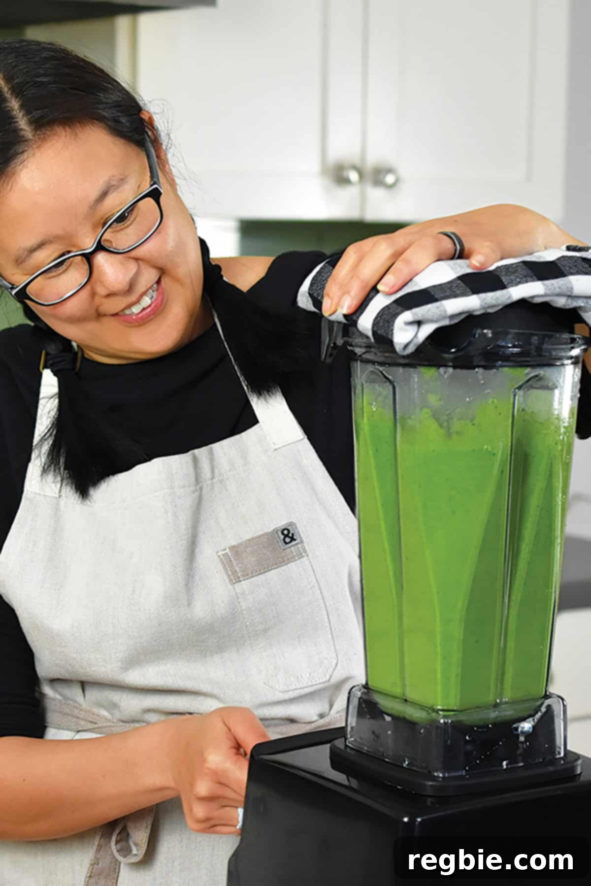 A smiling woman wearing glasses is expertly blending a rich, bright green soup in a high-speed Vitamix blender, demonstrating the ease of preparation.