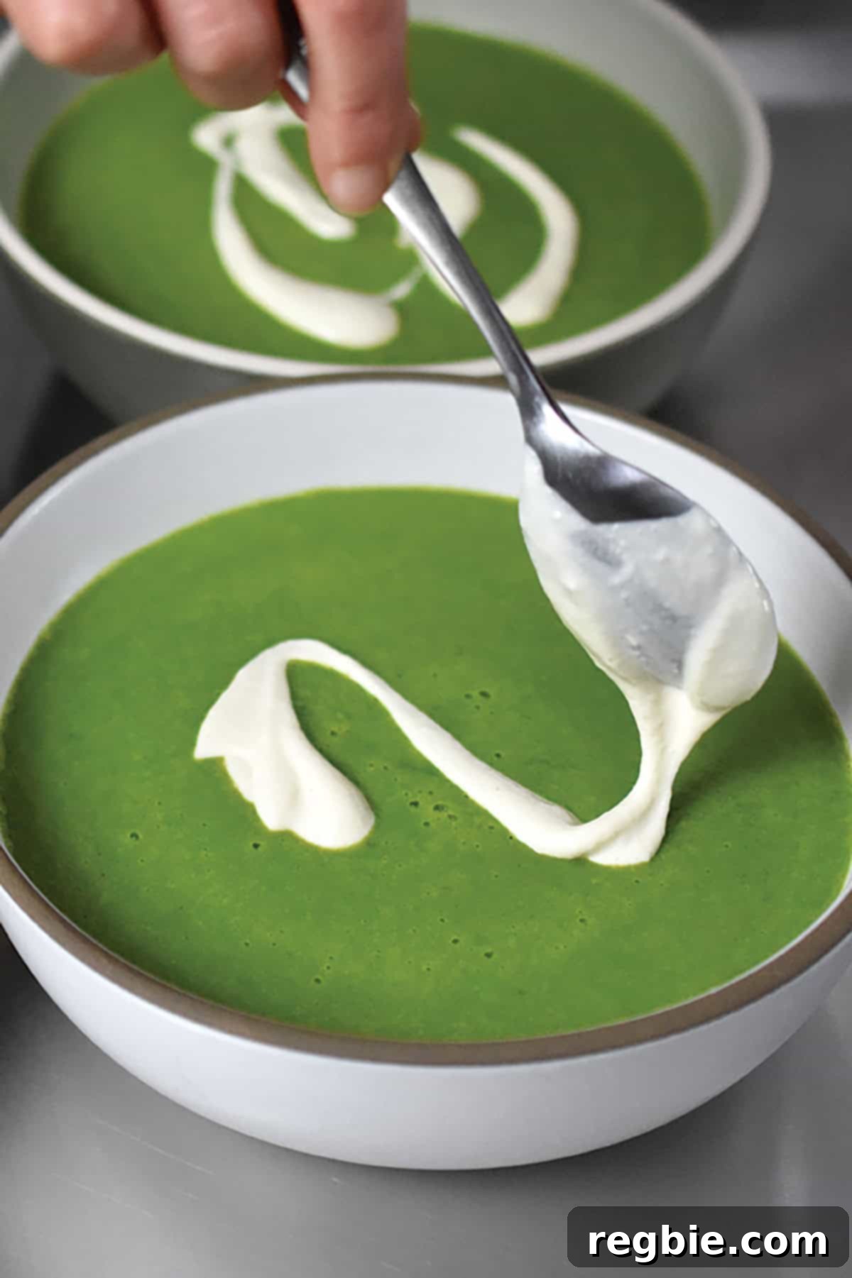 A dollop of creamy, dairy-free cashew cream being artfully added to the top of a bowl of vibrantly pureed green soup, ready to be served.