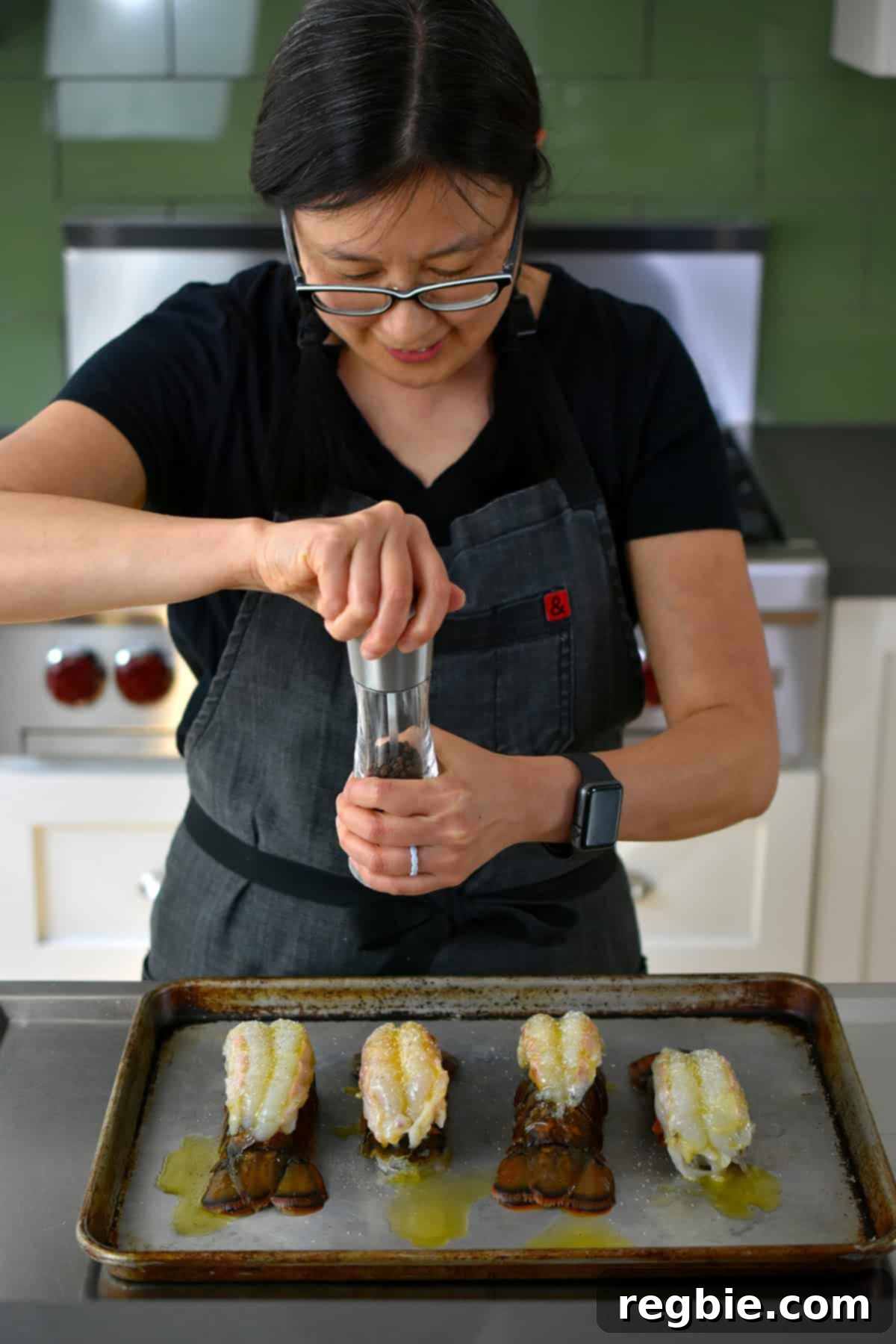 An Asian woman in glasses is adding freshly ground black pepper on top of raw butterflied lobster tails in a rimmed baking sheet.