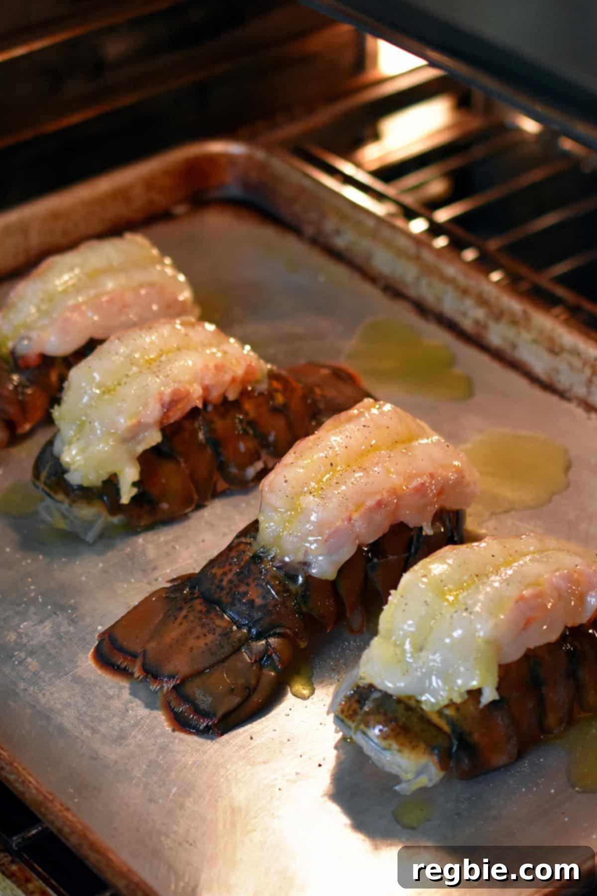 Four butterflied lobster tails on a rimmed baking sheet are baking in the oven.