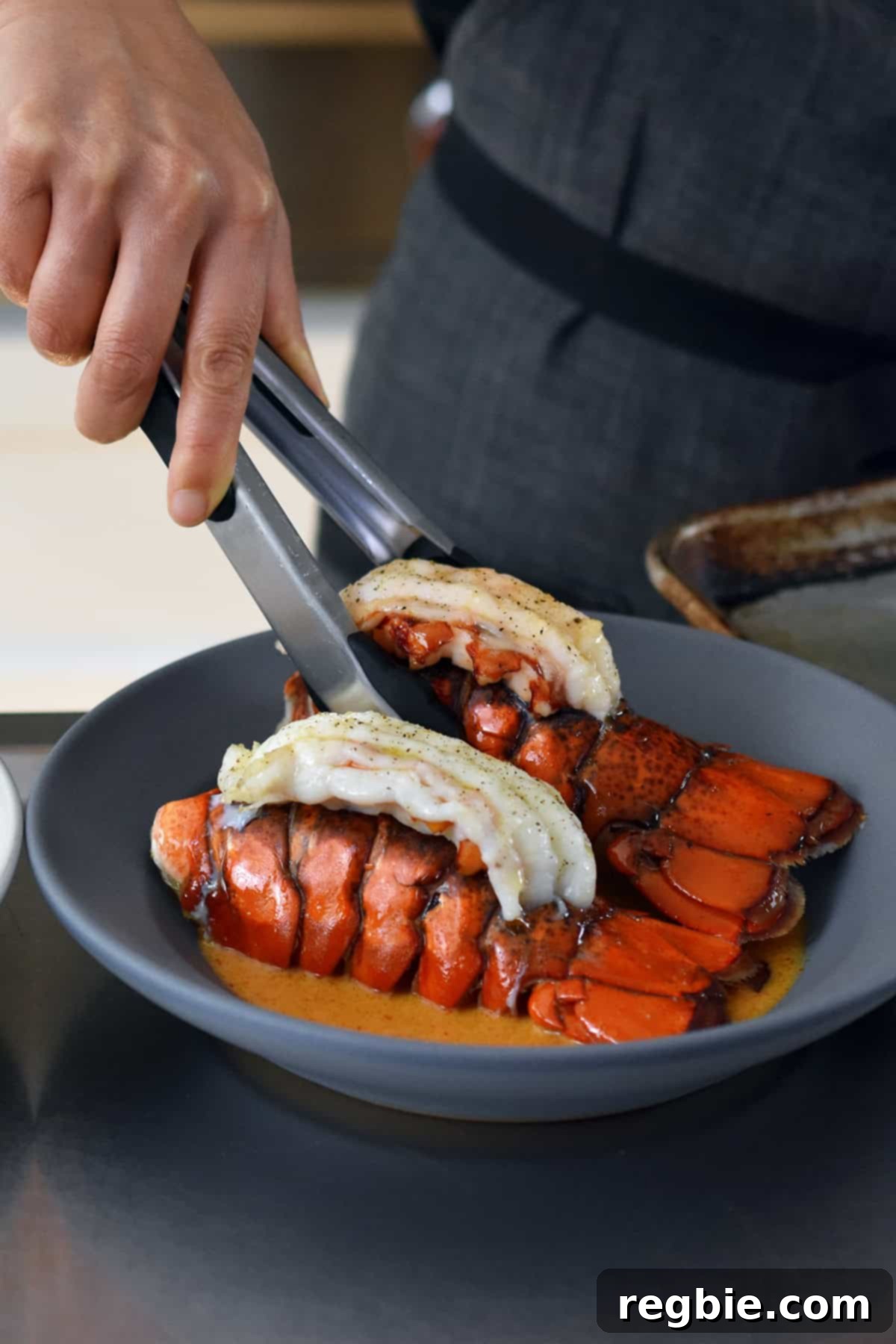 Using a pair of tongs to place two baked lobster tails in a shallow gray bowl filled with homemade Thai red curry sauce.