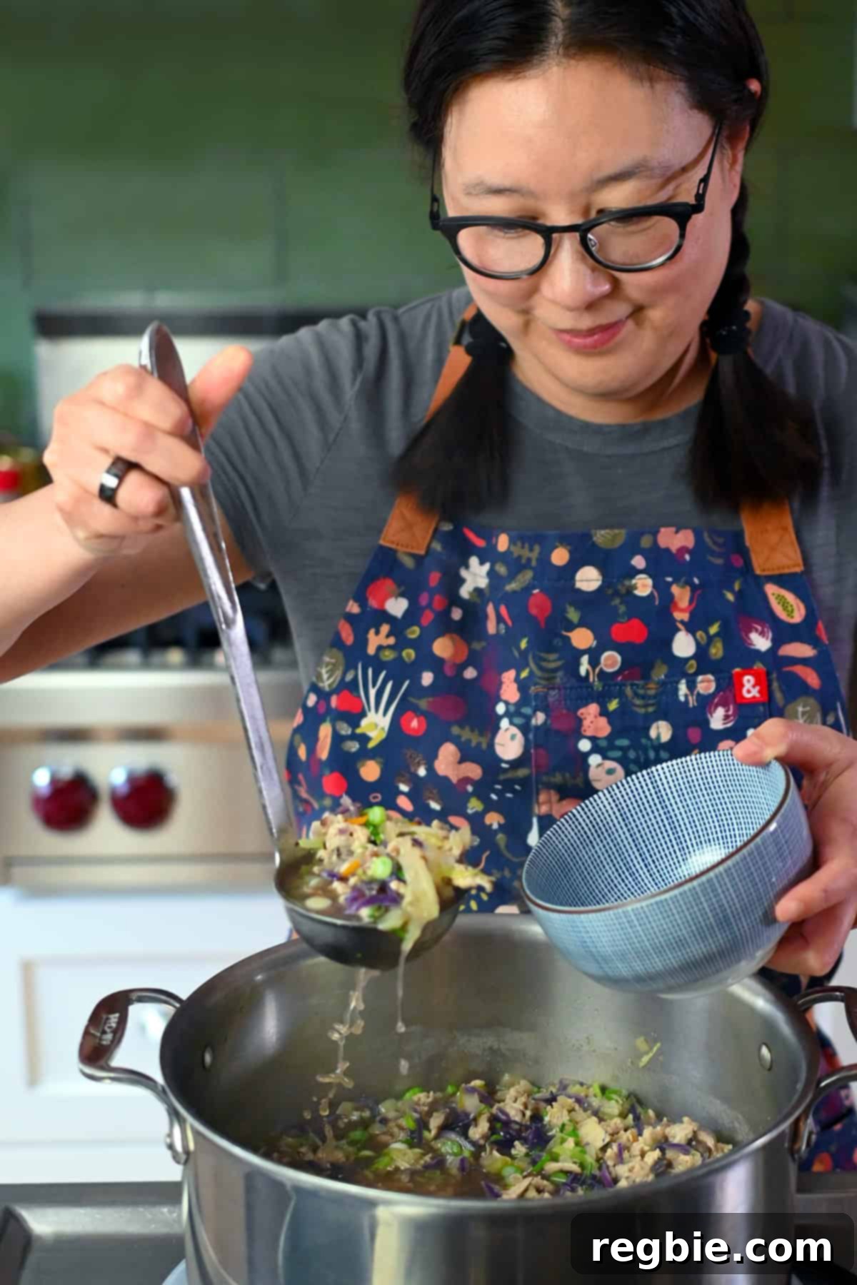 A smiling Asian woman in glasses and braids is scooping Asian chicken and cabbage soup into a serving bowl.