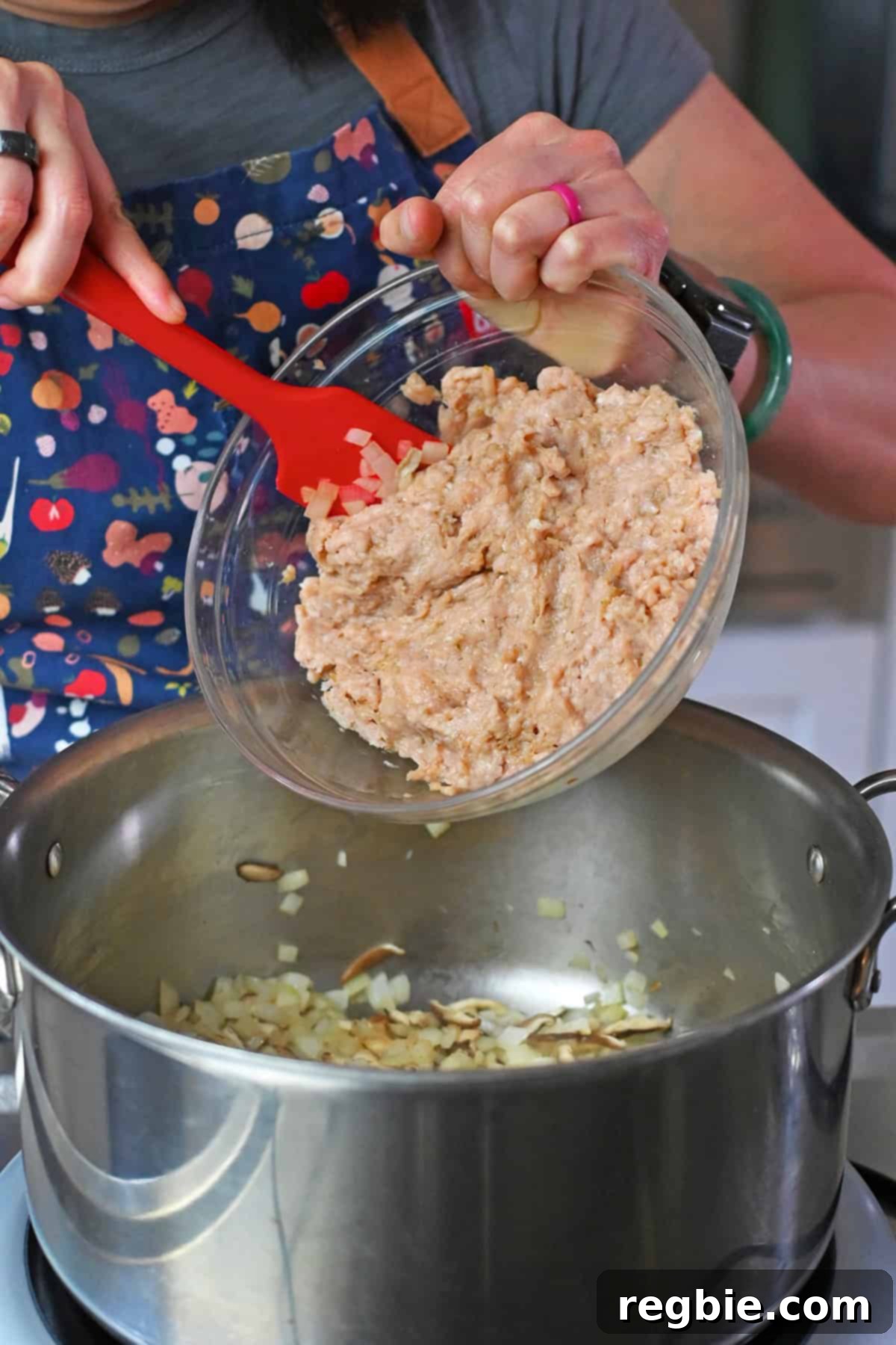 Adding the seasoned raw ground chicken into a stockpot filled with sautéed diced onions and sliced shiitake mushrooms.