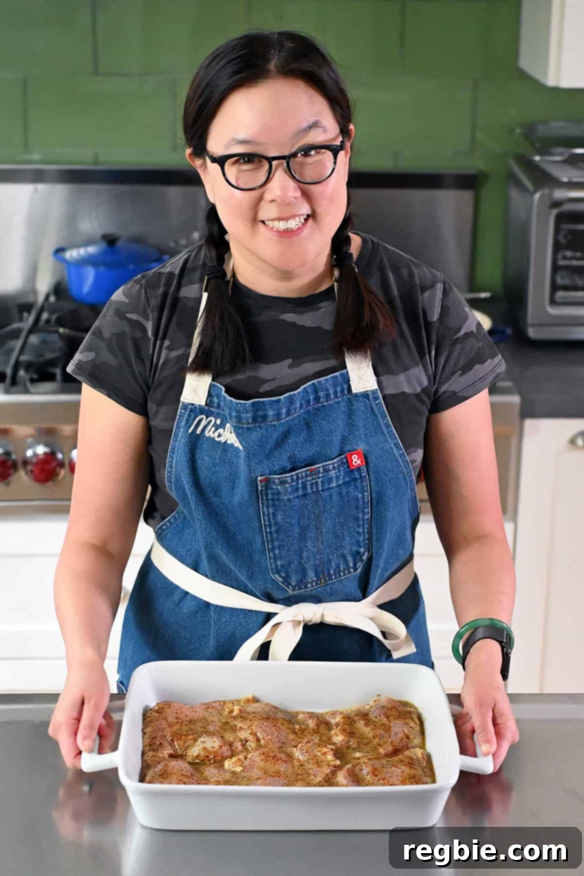 A smiling Asian woman in glasses and braids is holding a white casserole dish filled with honey mustard chicken thighs ready for the oven.
