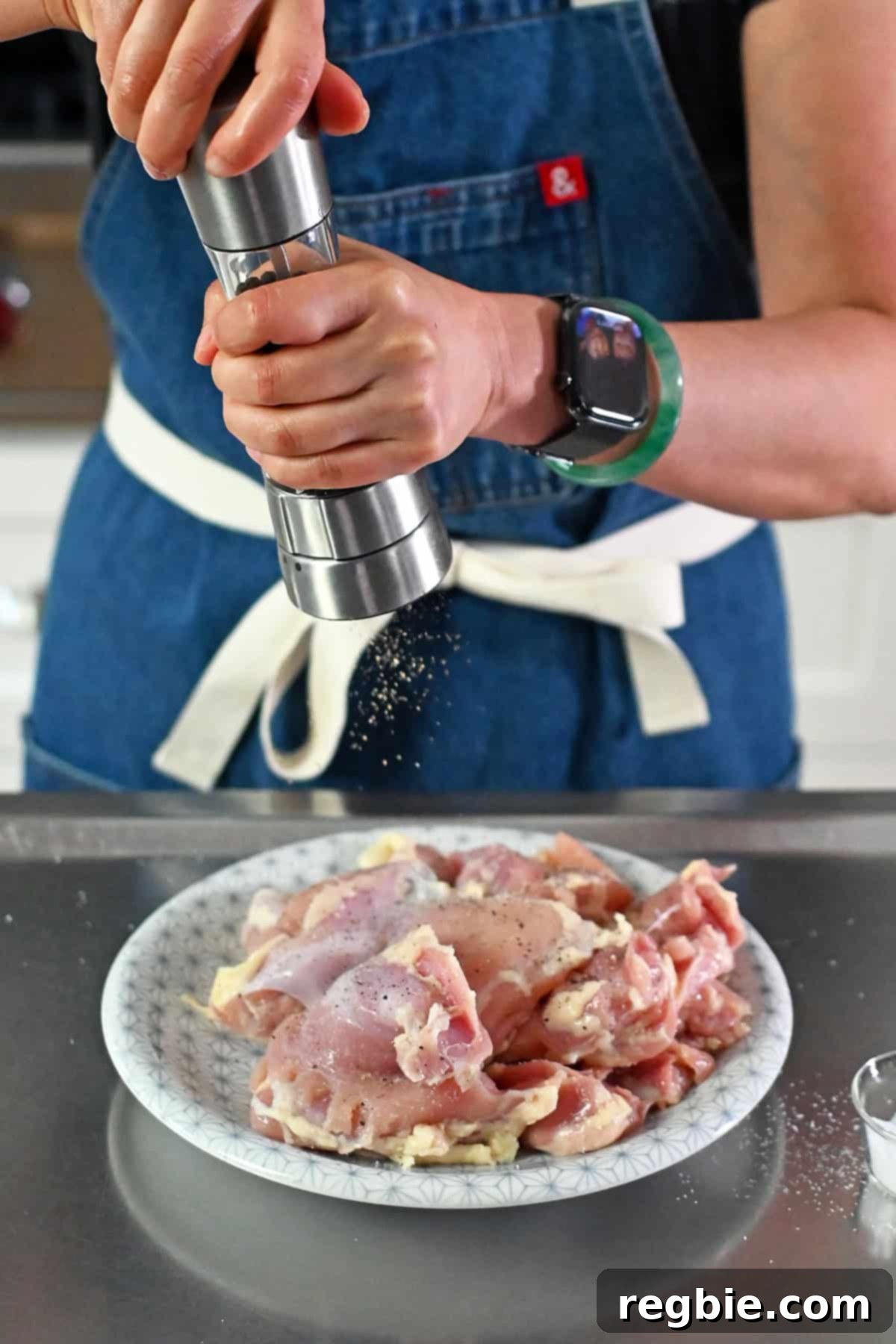 A person in a blue apron is adding freshly cracked black pepper to a plate of boneless and skinless chicken thighs.