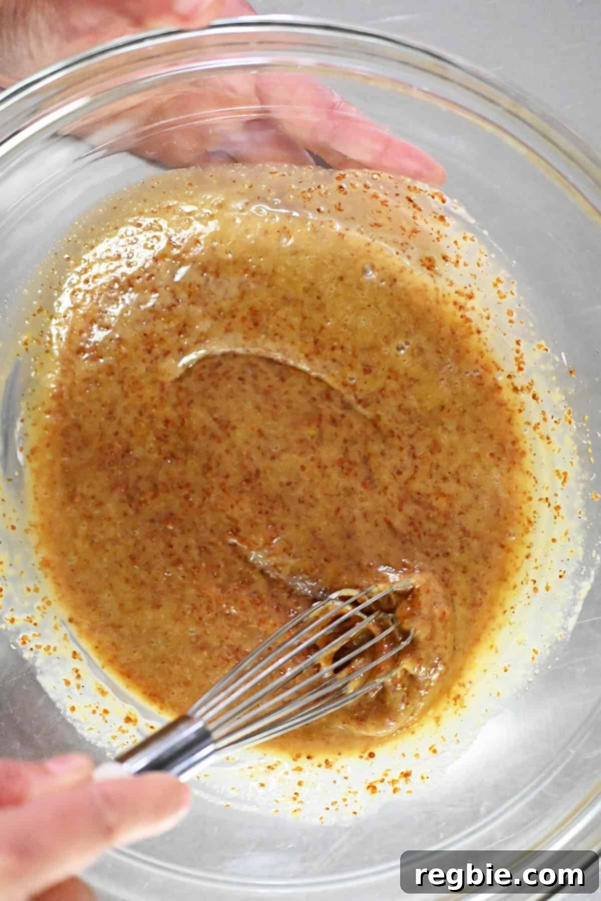 An overhead shot of a glass mixing bowl filled with honey mustard marinade being mixed with a small metal whisk.