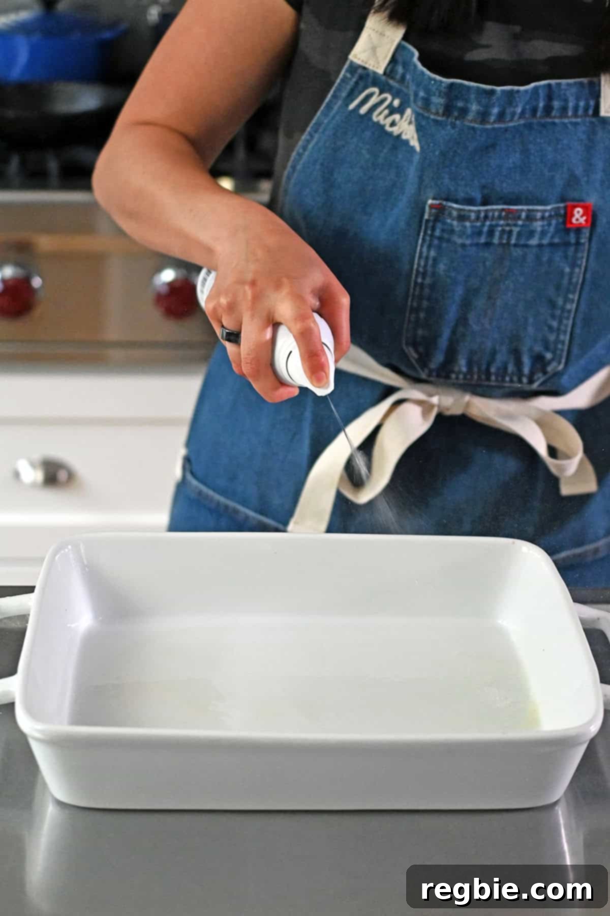 A person in a blue apron is spraying the inside of a large white rectangular baking dish with avocado spray.