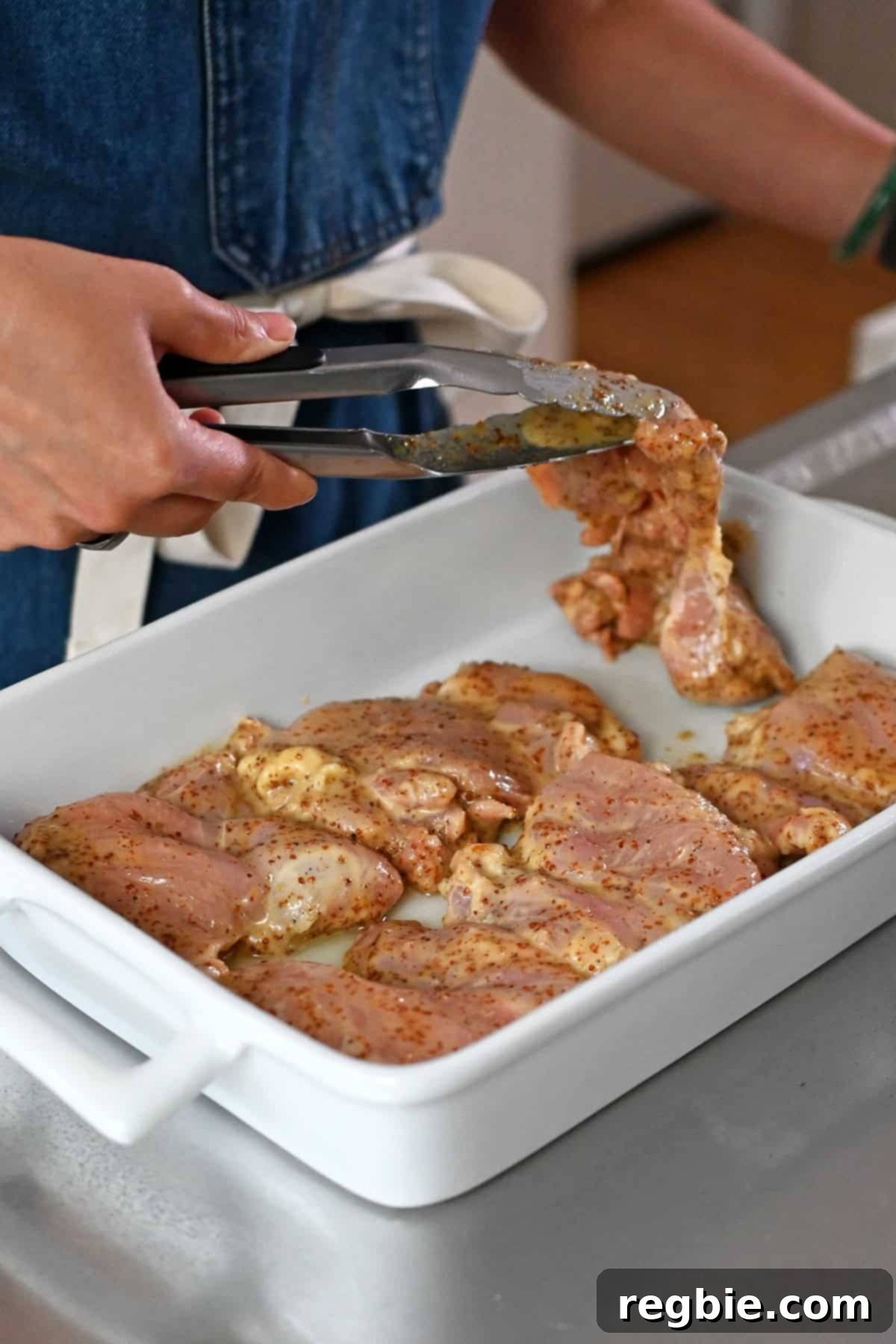 A person in a blue apron is placing the marinated raw honey mustard chicken in a single layer in a greased baking dish.
