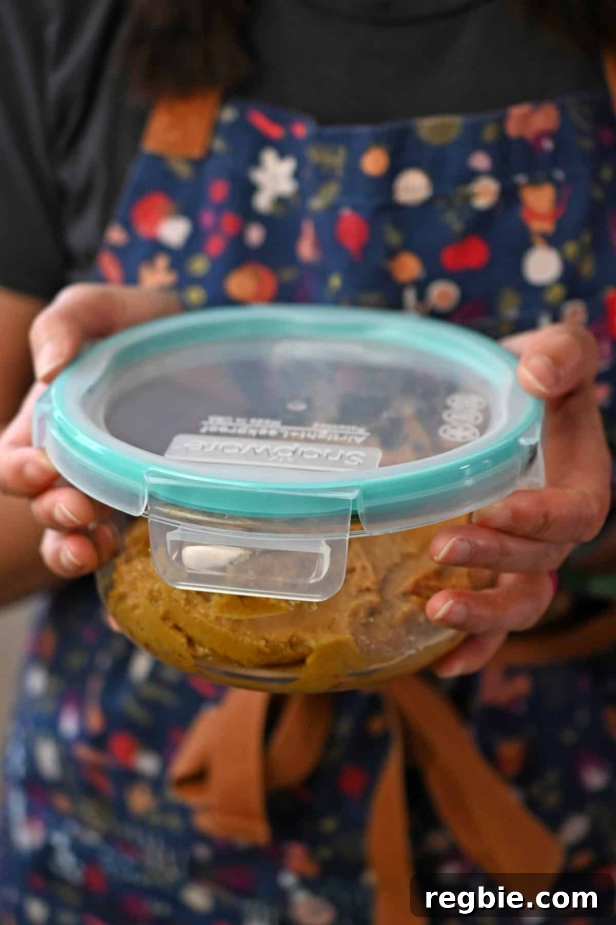 A person in a blue apron is holding a sealed round glass container filled with chilled and raw pumpkin spice snickerdoodle cookie dough.