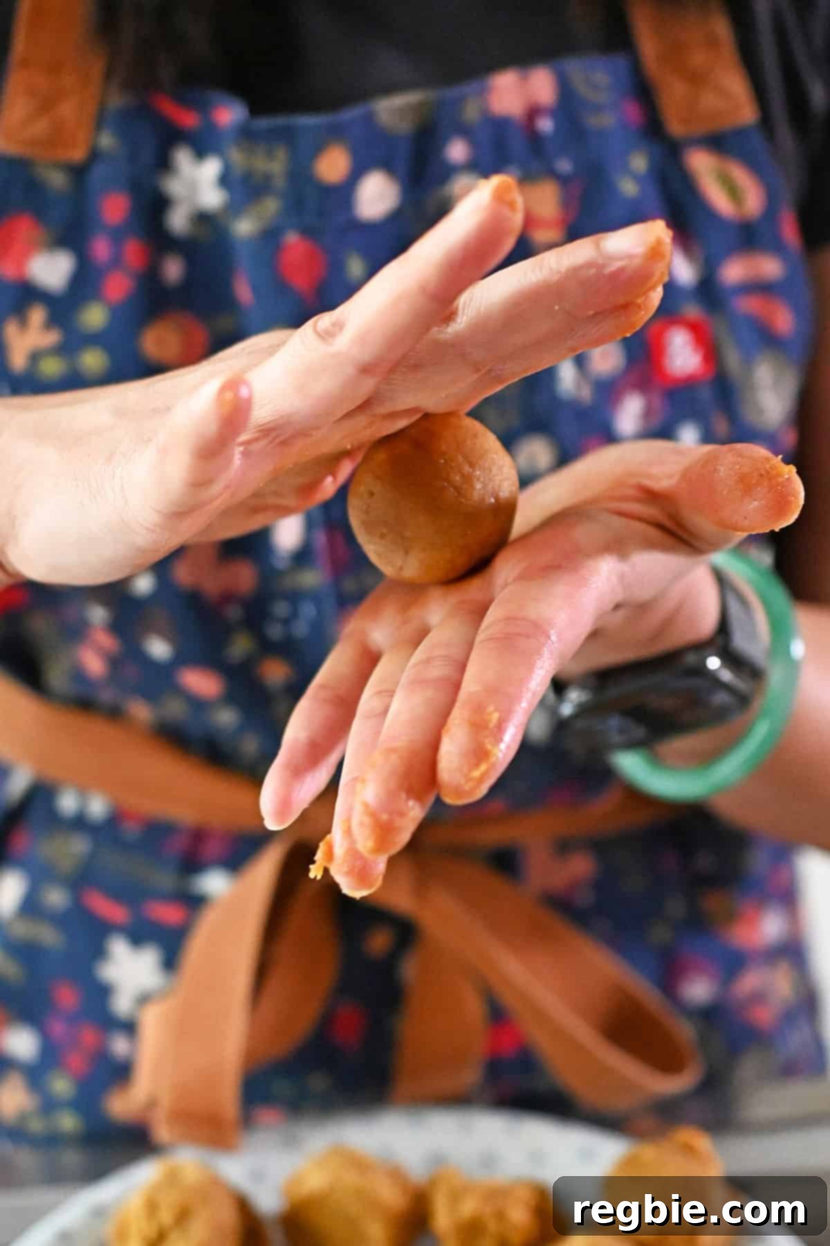 A person in a blue apron is rolling the raw pumpkin snickerdoodle cookie dough balls into smooth balls between their hands.