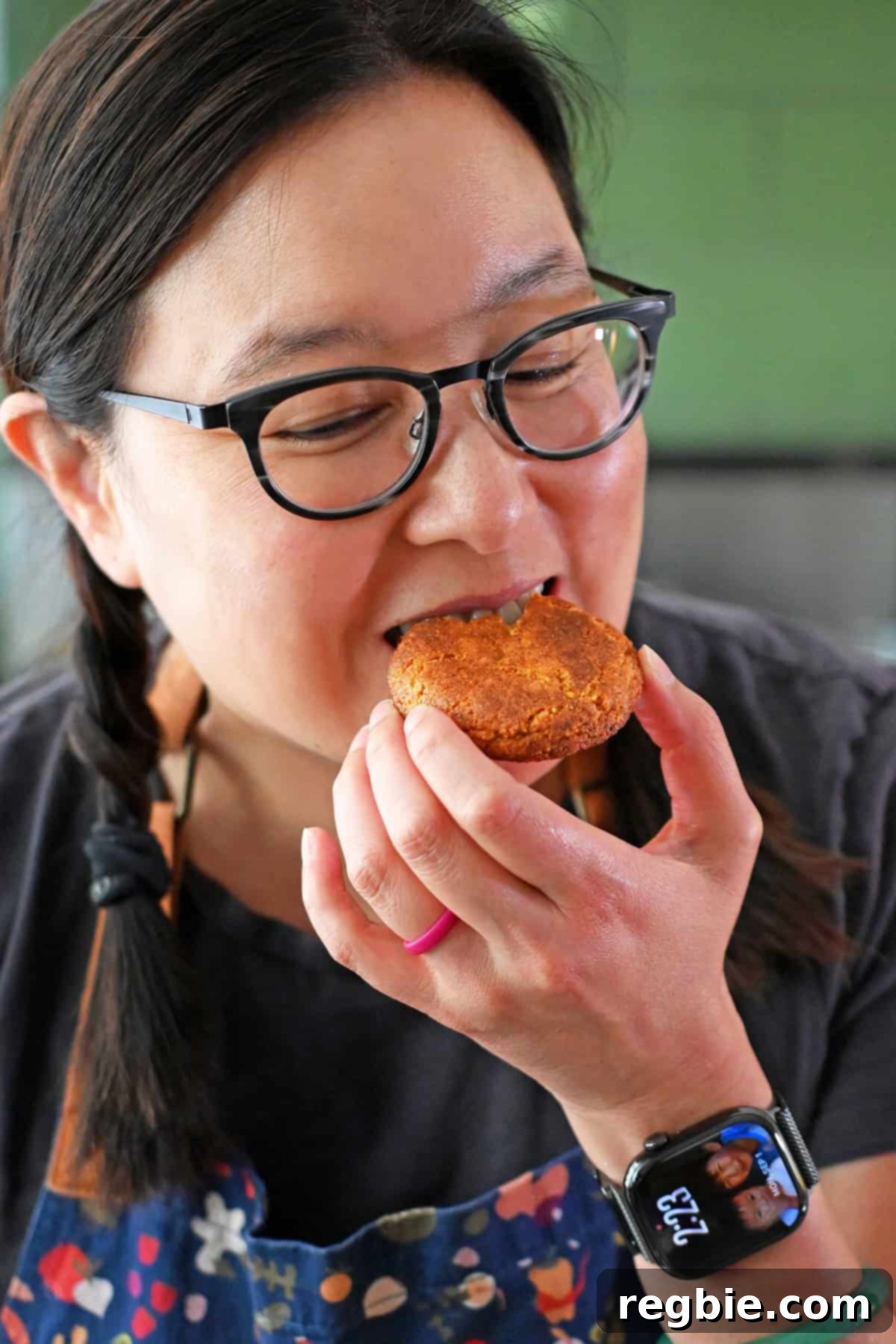 An Asian woman in glasses is biting into a freshly baked pumpkin snickerdoodle cookie.