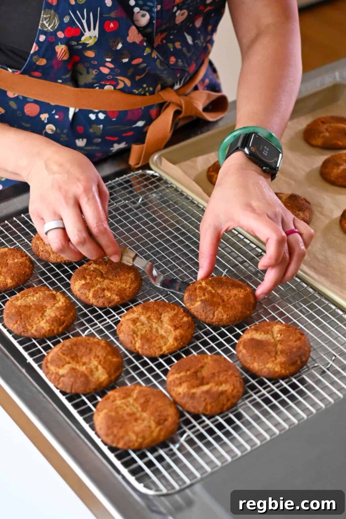 A person in a blue apron is transferring freshly baked pumpkin snickerdoodle cookies from a baking sheet to a cooling rack with an offset spatula.