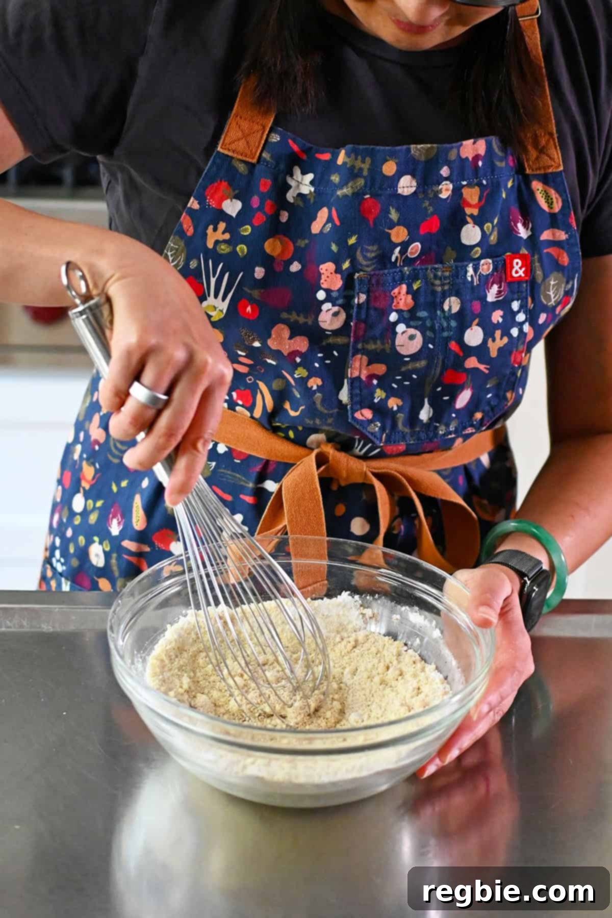 A woman in a blue apron is whisking together the dry ingredients to make paleo and gluten free pumpkin snickerdoodle in a glass mixing bowl.