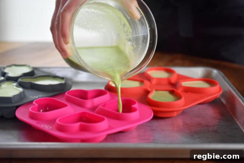 The green matcha coconut mixture being carefully poured into various silicone molds on a baking tray.